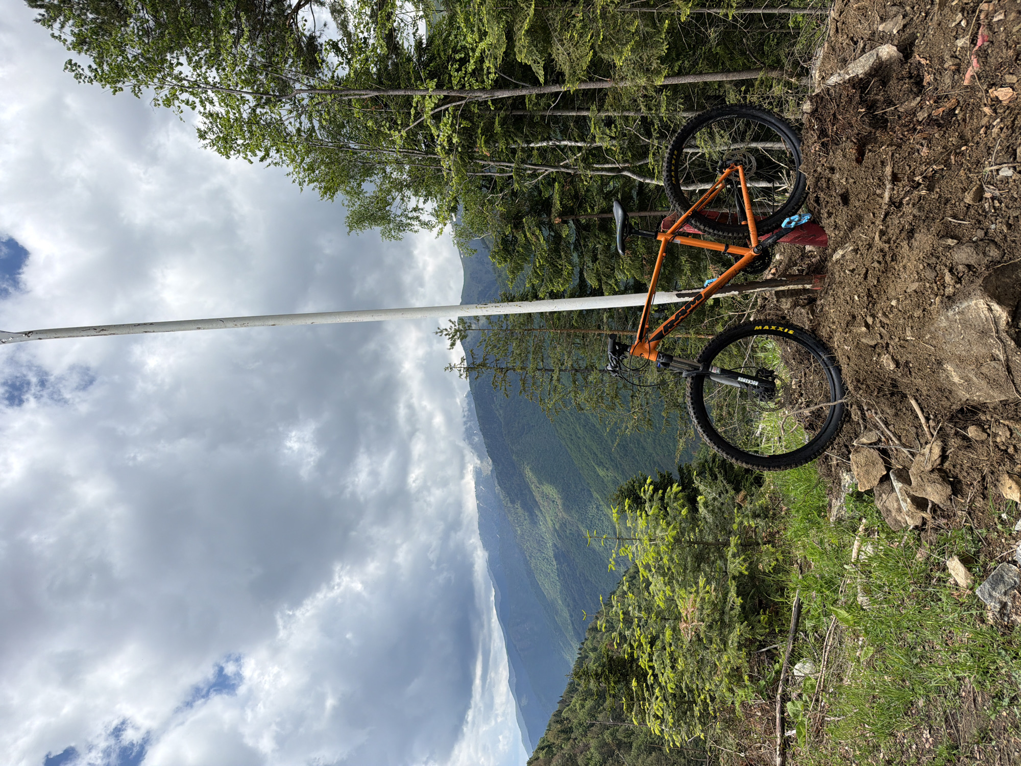 An orange mountain bike is positioned on a dirt ledge next to green foliage, set against a backdrop of mountainous terrain and a cloudy sky. A tall, white pole is visible in the foreground. The location is near Рилски манастир, Bulgaria.