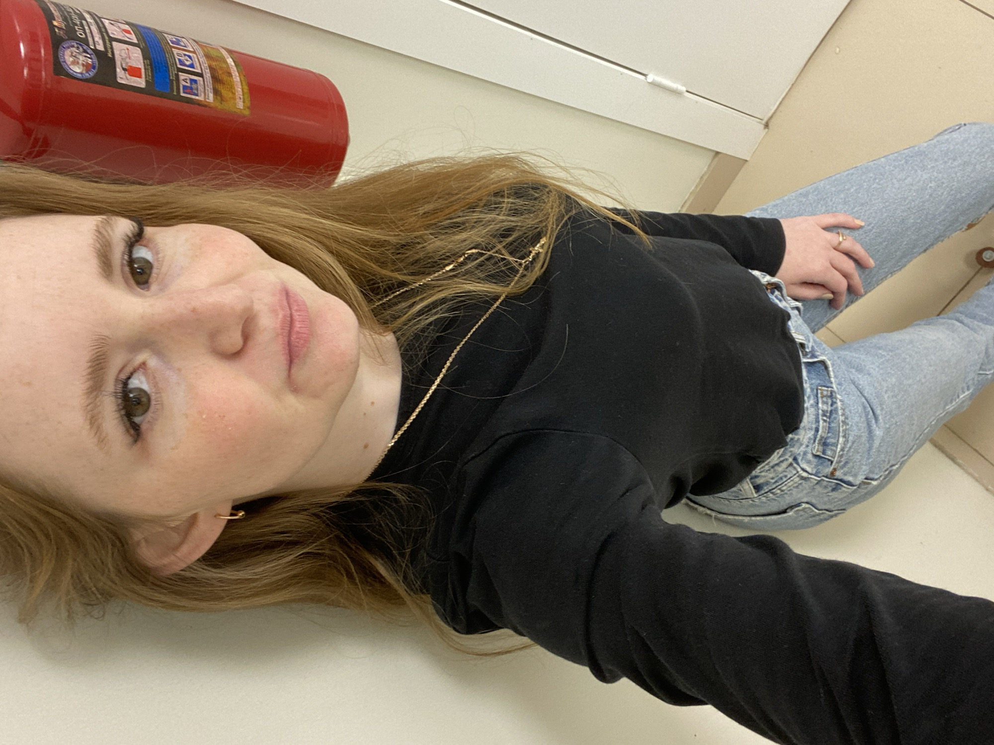A young woman is captured in a selfie, lying on a light-colored floor and looking directly at the camera with a slight smile. She has long, reddish-brown hair, light brown eyes, and a fair complexion with visible freckles. She is dressed casually in a black long-sleeved top and light blue jeans. Her accessories include a thin gold chain necklace, a small gold hoop earring in her left ear, and a gold ring on her right hand.
The setting appears to be an indoor space, likely a hallway or office, with plain light-colored walls and floor tiles. A red fire extinguisher is visible behind her head, positioned horizontally. The fire extinguisher features a label with Cyrillic text, including "ОП-10", and pictograms illustrating different fire classes (A, B, C, E) and usage instructions. The image suggests a relaxed, casual moment captured indoors in Chaikovsky, Russia, with no specific time of day or weather discernible due to the indoor lighting.