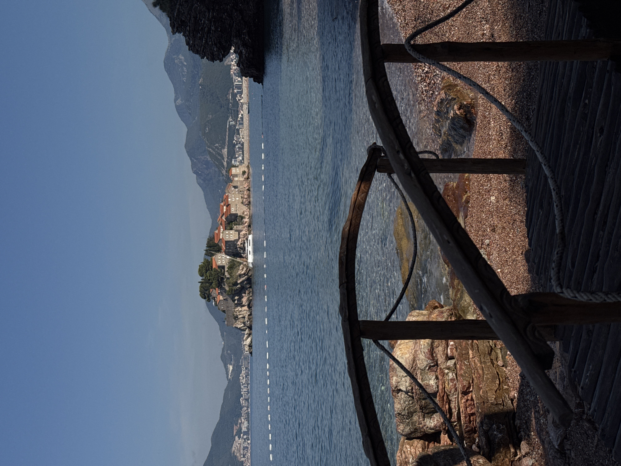 The image captures a scenic view of the island of Sveti Stefan, a fortified island town located in Montenegro. The foreground features a weathered wooden walkway and railing, with thick ropes acting as a barrier. Beyond the walkway, a pebble beach transitions into the clear blue waters of the Adriatic Sea. In the middle ground, the island of Sveti Stefan rises from the sea, characterized by its stone buildings with red-tiled roofs, nestled amidst lush greenery. A white yacht is anchored near the island. The background is dominated by rolling green mountains under a clear blue sky. The lighting suggests it is daytime, with the sun casting shadows on the beach and walkway.