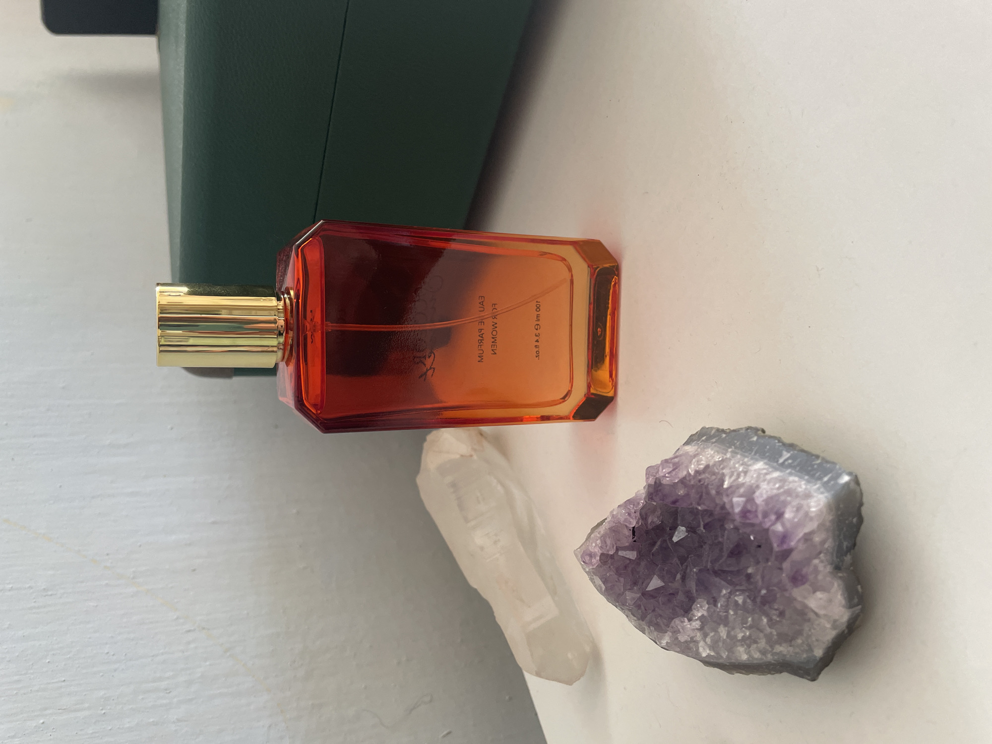 The image depicts a vanity scene, likely in a domestic setting, featuring a perfume bottle with a gradient of orange and red hues, topped with a gold cap. To the left of the perfume bottle are two crystals: a clear, elongated quartz crystal and a cluster of amethyst. In the background, a dark green box is visible. The lighting suggests an indoor environment, possibly near a window or under artificial light. The image does not contain any information or visual cues that would allow for its location to be determined as Kopeysk, Russia, or any specific city.