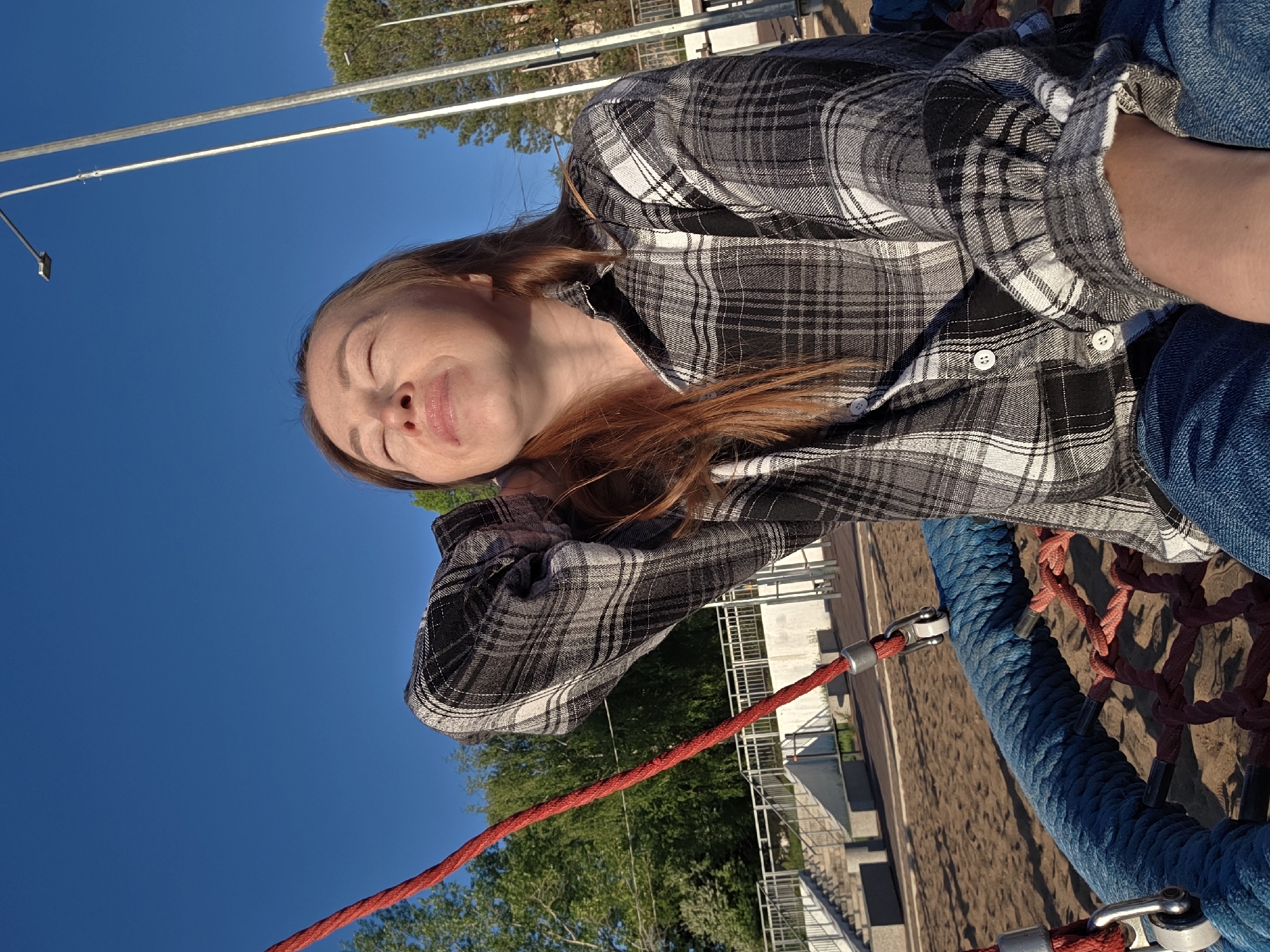 The image shows a woman with long red hair wearing a grey and black plaid shirt and blue jeans. She is lying on a blue rope swing with red rope attachments, closing her eyes and making a face. The background includes a clear blue sky, green trees, and a building or structure with white railings. The photo appears to have been taken in the city of Chaikovsky, Russia, judging by the presence of such structures.