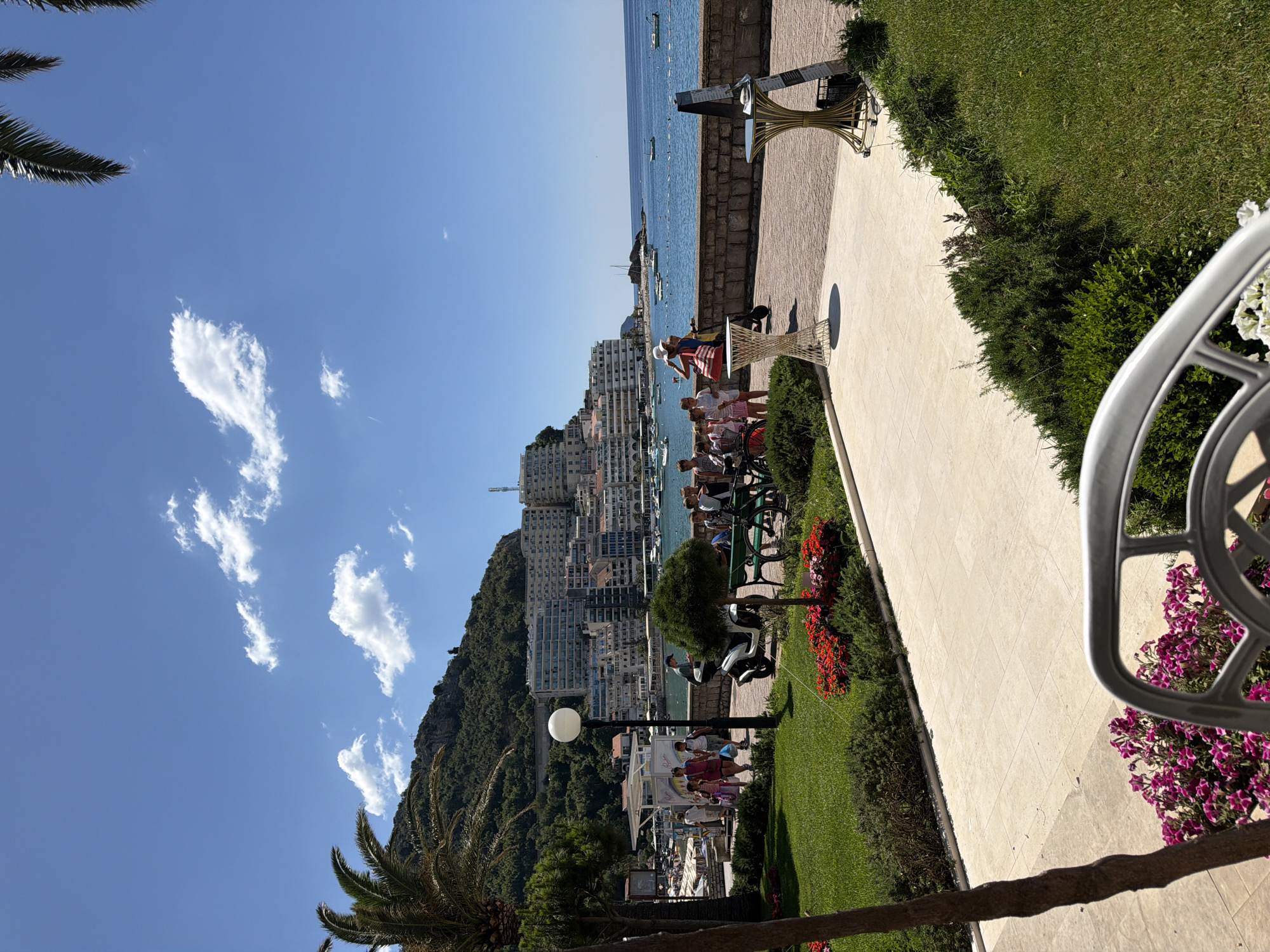 In an outdoor scene under a clear blue sky with scattered white clouds, a paved walkway borders a manicured green lawn with flowerbeds. In the background, a body of water with several boats is visible, beyond which lies a hillside city with numerous buildings and lush green vegetation. On the right side of the walkway, a tall stone wall separates the paved area from the water. In the foreground, a white metal chair is partially visible.
Several people are present in the scene. A group of children and adults are gathered near a bench and a scooter on the walkway. Another group of people is walking further down the walkway towards the city. A person on a scooter is riding on the walkway. A couple of decorative tables are placed on the walkway. Palm trees are seen on the left and right sides of the image. The overall ambiance is that of a sunny day in a coastal city.