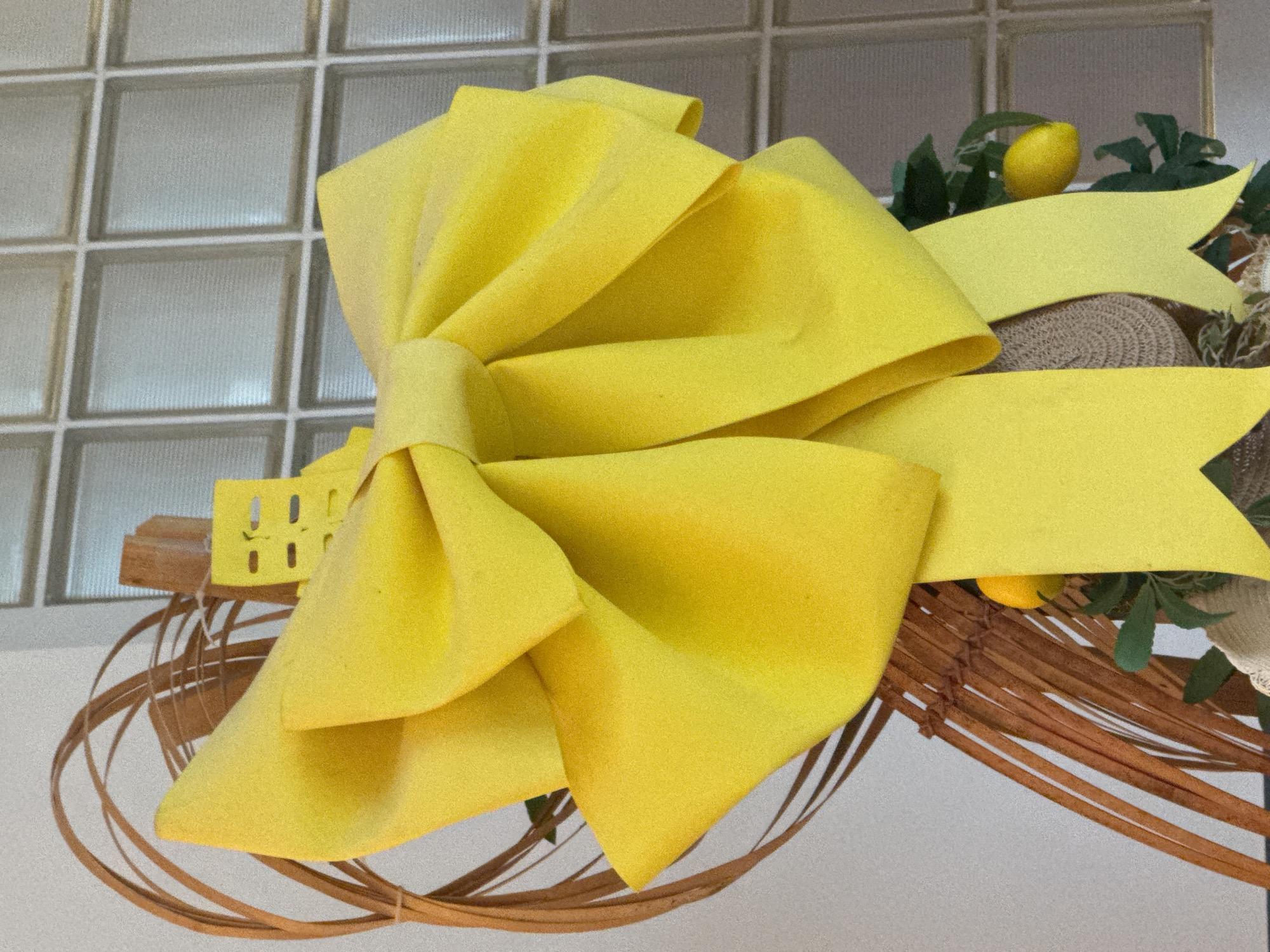 A close-up shot shows a large, bright yellow bow dominating the scene. It appears to be made of a stiff material, possibly felt or thick fabric, with a textured surface. The bow is intricately folded and tied, creating a multi-layered effect.
Behind the bow, there's a wreath-like structure made of intertwined brown twigs and branches. Small lemons and green leaves are interspersed within the wreath, adding a touch of nature and freshness. Additionally, a woven mat is visible.
The backdrop features a grid-like frosted glass panel.
There's no evidence to suggest that this photo was taken in Guilin, China.
