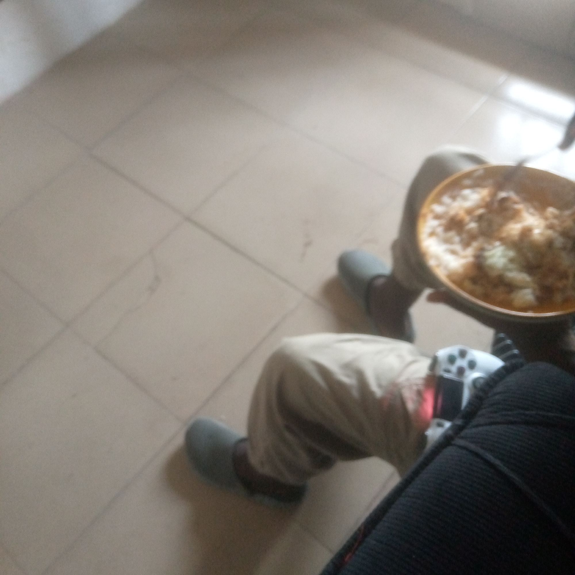 In an indoor setting, a person is seated on a tiled floor. They are wearing light-colored pants and what appear to be grey slippers. The person is holding a bowl of food, which seems to be a rice dish. Next to them, on the floor, is a white video game controller. The lighting suggests an indoor environment, possibly a room with natural light coming from a window or opening. The background is out of focus but shows more of the tiled floor.