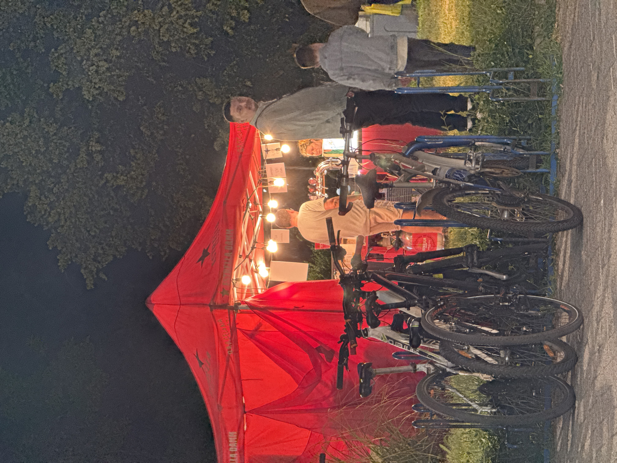 The image depicts an outdoor scene at night, likely in the city of Burgas, Bulgaria, near the Sea Garden. In the foreground, several bicycles are parked in a rack. A red tent or canopy, illuminated by string lights, stands in the background. Behind the tent, a bar area is visible with taps. A man with short dark hair and a light-colored jacket is standing in front of the bar, looking towards the camera. To his right, a younger person in a grey jacket is also visible. Another man in a light-colored shirt and shorts is standing at the bar. The overall atmosphere suggests an event or gathering at a food or drink stall.