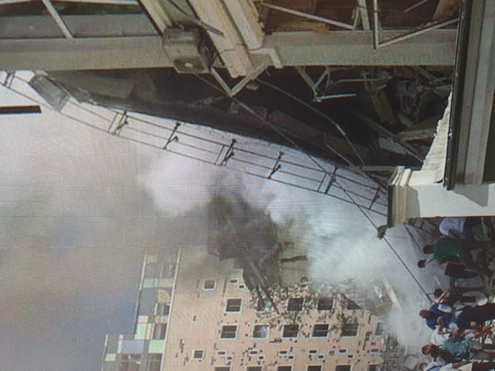 This image depicts the aftermath of an explosion at the Central Research Institute of Mechanical Engineering in Tashkent, Uzbekistan. A building on the right, with a white facade and dark, angular metal structures, shows significant structural damage, with debris and a large, curved metal element exposed. Smoke and dust billow from this damaged structure, obscuring the background.
To the left, another building, lighter in color and with numerous dark rectangular windows, also appears to have sustained damage, with some sections appearing partially collapsed or riddled with holes. Debris is visible in the foreground and around the base of this building.
In the lower portion of the image, several people are gathered, some appearing to be observing the scene or possibly involved in rescue or assessment efforts. They are dressed in casual clothing, including t-shirts and shorts. The environment is urban, characterized by the presence of these large buildings and the visible destruction. The overall scene is one of devastation and disarray.