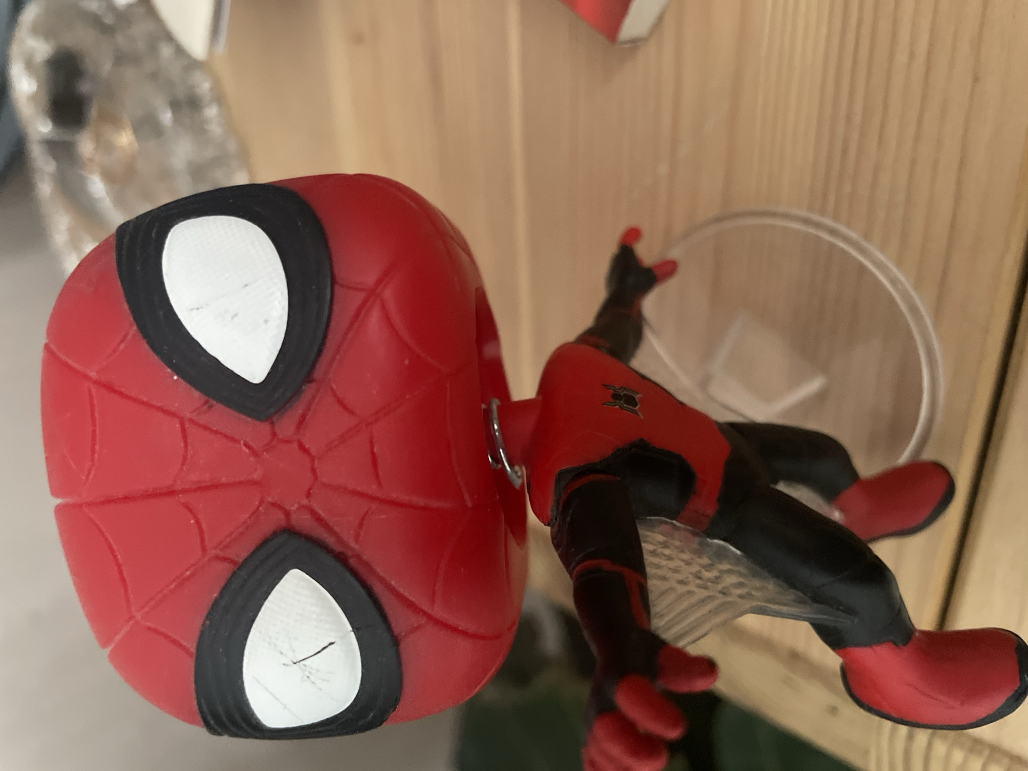 The image shows a close-up of a Spiderman action figure, likely a Funko Pop, in a red and black suit. The figure is positioned horizontally, with its large, red head with white eye lenses dominating the left side of the frame. The intricate red webbing pattern on the head is clearly visible. The figure's body is connected to its head by a metal post. The right side of the image shows the figure's torso, arms, and legs, also in red and black. The figure is supported by a clear plastic stand, which is mostly out of focus. The background consists of a light-colored wooden surface, possibly shelving, and some blurred objects in the upper left corner, one of which appears to be a clear plastic container or bottle. The lighting suggests an indoor setting, and there are no indications of weather or time of day. No text is visible in the image. The overall impression is that of a display of collectibles.