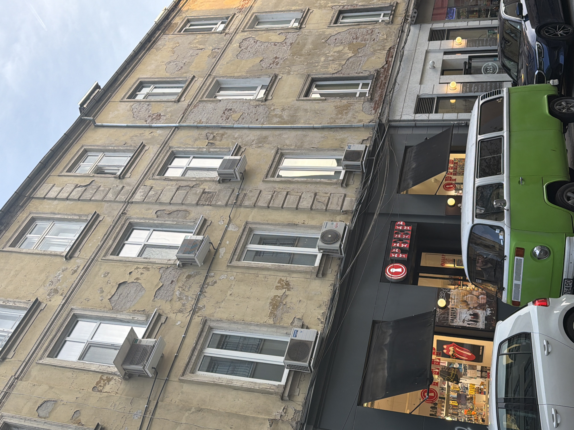 The image shows a multi-story building with a weathered facade, featuring multiple windows and several air conditioning units attached to the exterior walls. Adjacent to the building is a modern structure with storefronts, visible through dark awnings. A bright green vintage Volkswagen bus is parked in front of the modern building, with other vehicles partially visible, including a white car. The sky is overcast. The location is Unknown, Unknown.