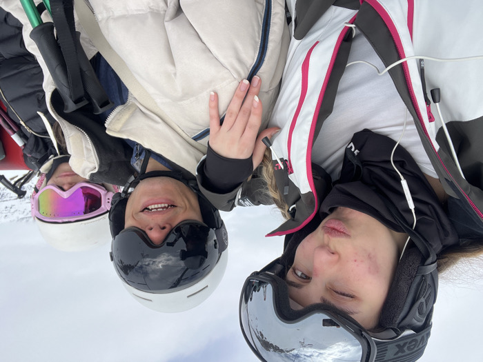 Three young individuals are captured in a selfie-style, close-up shot, appearing to be on a ski slope or lift. The original image is rotated 90 degrees clockwise, showing the subjects looking upwards towards the camera.
From left to right:
The leftmost person wears a white ski helmet and bright pink reflective goggles, with a dark jacket partially visible. Their face is mostly obscured by the goggles, but their mouth is visible.
The central person wears a white ski helmet and dark ski goggles, smiling broadly and showing their teeth. A hand with manicured nails (French tips) rests near their shoulder. They are wearing a light beige or tan ski jacket.
The rightmost person wears a black ski helmet and large mirrored ski goggles, with the brand "UVEX" visible on the goggle strap. They are making a playful face with slightly puckered lips and one eye partially closed. White wired earbuds are visible in their ears. They are wearing a distinctive white, black, and pink ski jacket.
Ski poles are partially visible on the far left of the image. The background is a bright, blurry white, indicative of a snowy environment or cloudy sky, consistent with a mountain resort setting like Val-d'Isère, France. The activity taking place is likely a break during skiing or snowboarding. The time of day is daylight, and the expressions suggest a cheerful and relaxed mood.