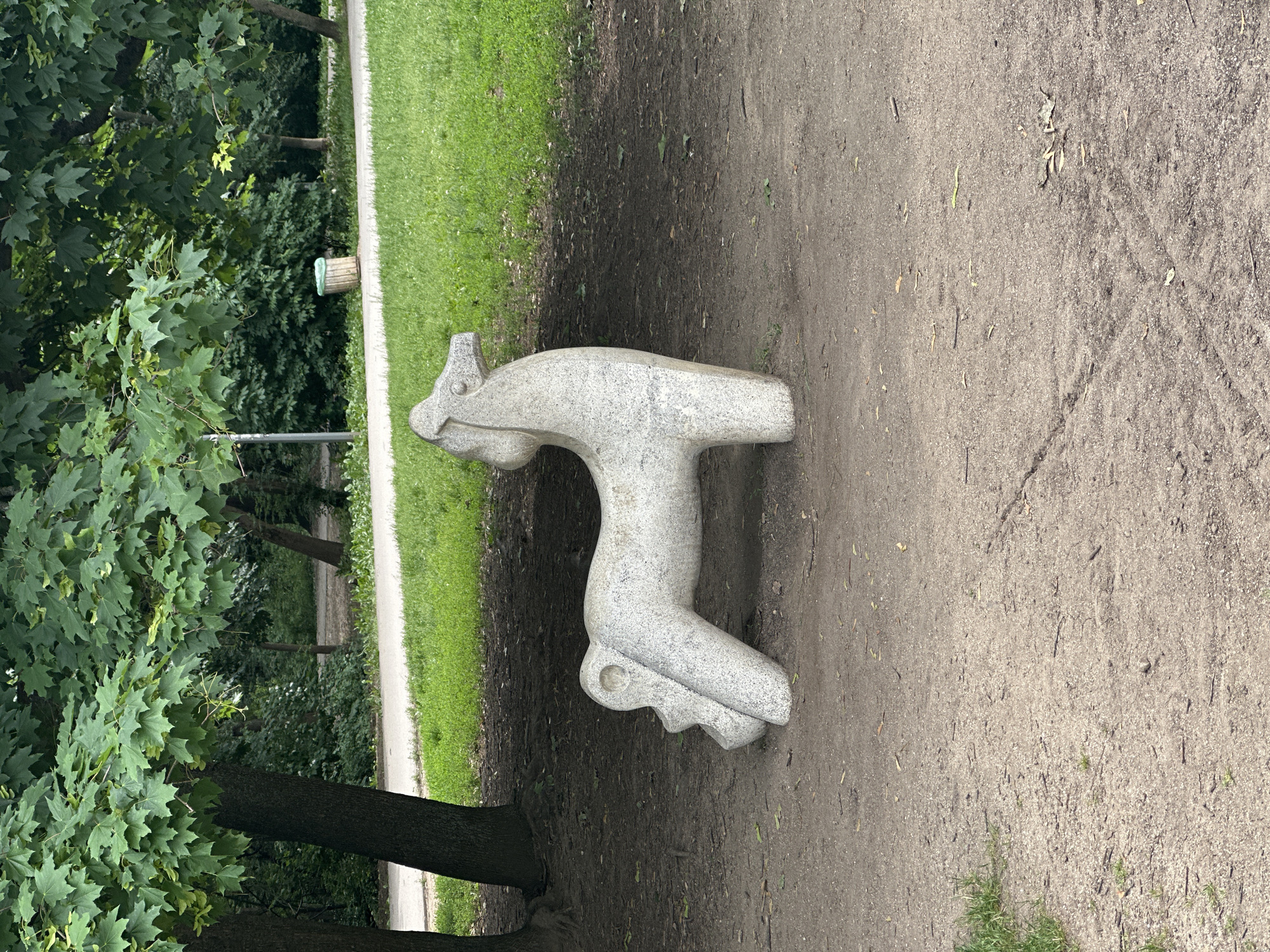The image depicts a light-colored, stone sculpture of a horse in a park-like setting. The horse sculpture is positioned on a dirt ground, with a grassy area and trees in the background. The horse is facing to the right, with its head lowered slightly. Its body is stylized and rounded, with simplified legs and tail. The texture of the sculpture appears to be rough and porous, characteristic of granite or a similar stone.
In the background, a paved path runs horizontally, bordered by lush green grass. Beyond the path, there are more trees and bushes, indicating a natural or landscaped outdoor environment. A trash bin with a light-colored body and a green rim is visible to the right of the path. The lighting suggests it is daytime, with some shadows cast on the ground, particularly around the base of the trees.
This sculpture is located in the Borisova Gradina park in Sofia, Bulgaria. It is part of the children's play area within the park.