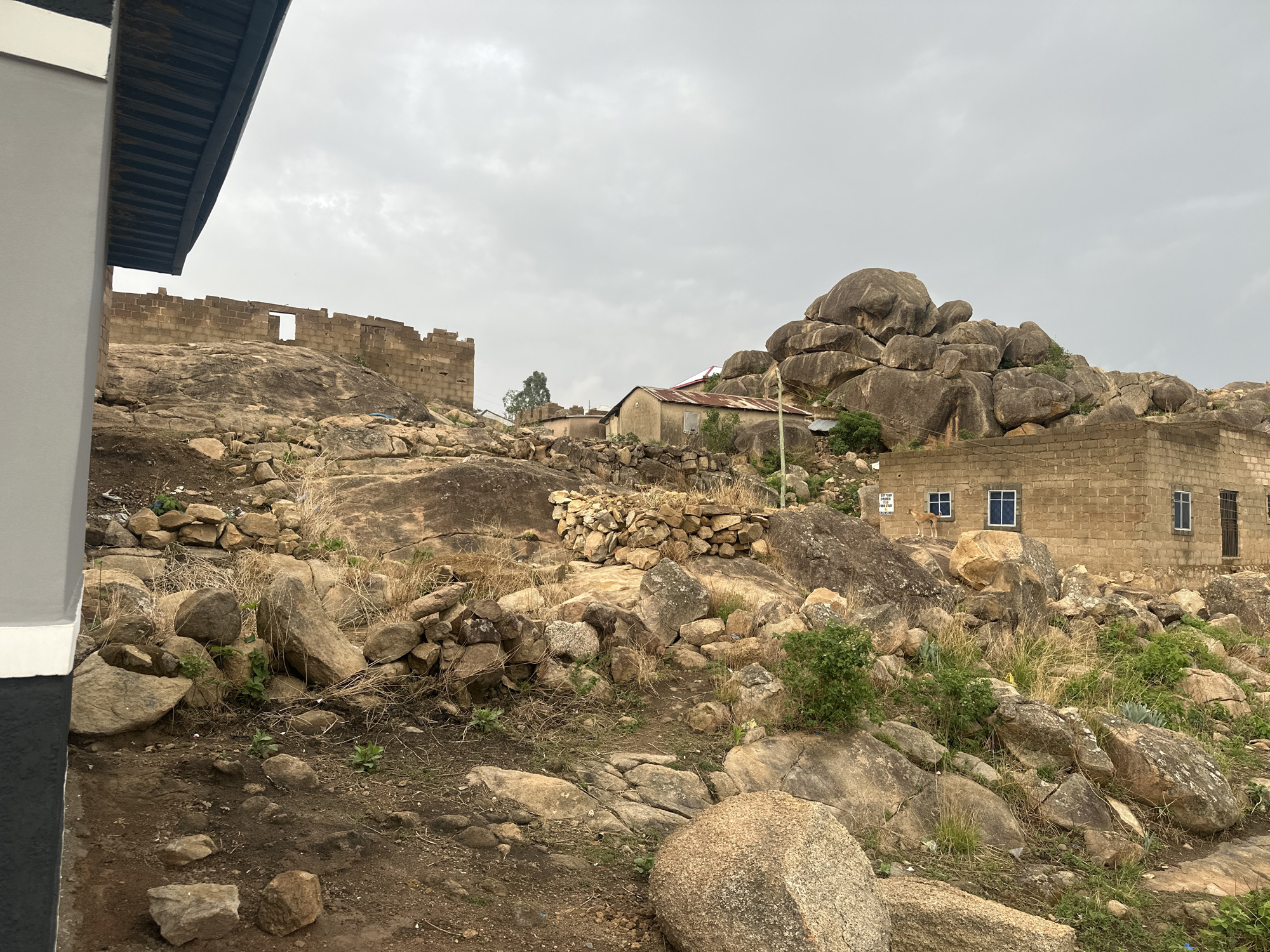 The image shows a rocky hillside in Jos, Nigeria. The foreground is dominated by large boulders and sparse vegetation. In the midground, there are a few buildings, including a concrete structure with windows, and a partially ruined brick building. A stack of piled-up rocks is visible on a slope. In the background, a cluster of large, rounded rocks is visible against a cloudy sky. No people are visible in the frame.