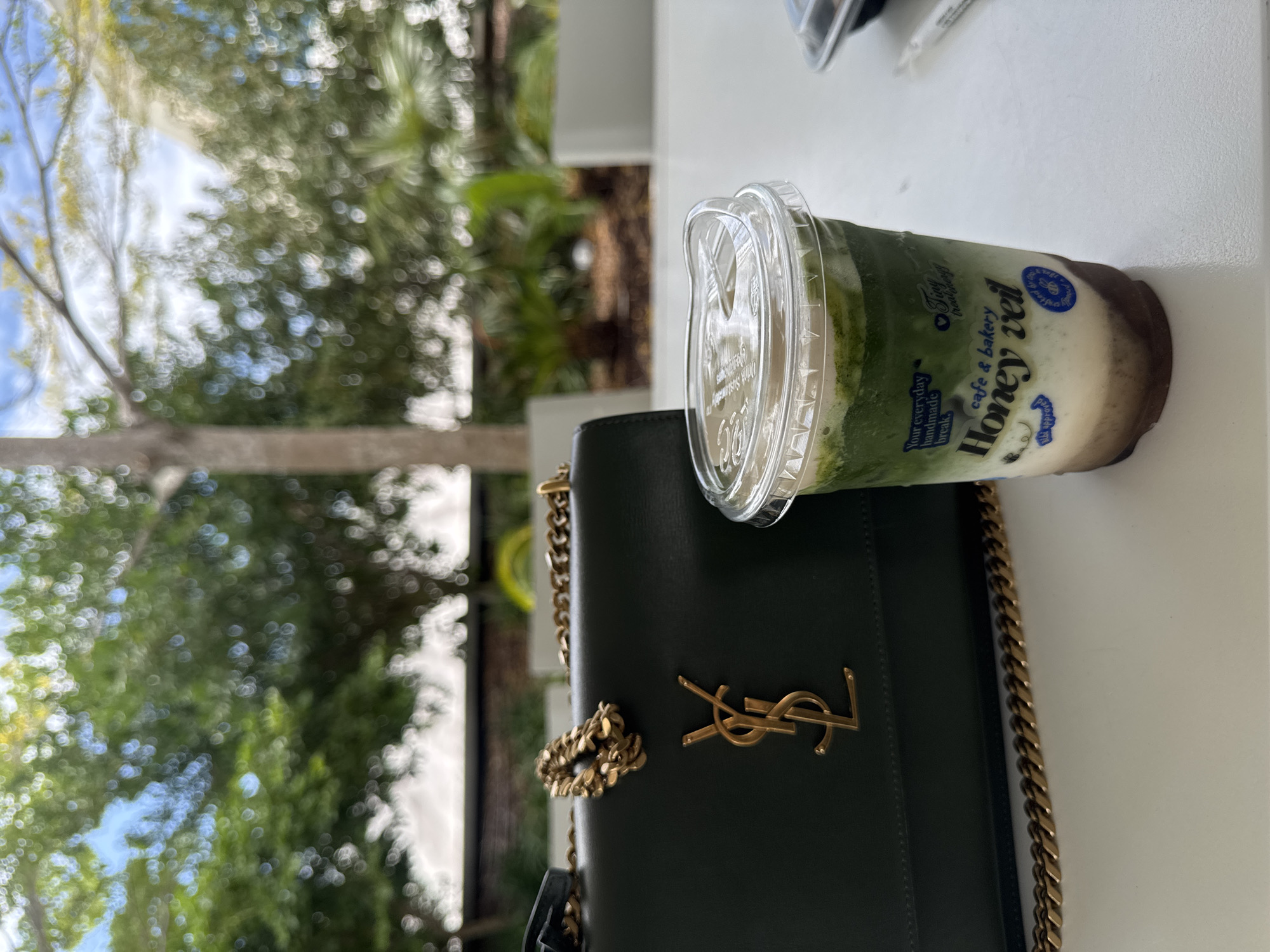 A dark green Yves Saint Laurent purse with a gold chain strap rests on a white table next to a clear plastic cup containing a layered green beverage. The purse features a prominent gold YSL logo. The beverage appears to be a matcha latte with layers of cream and possibly chocolate at the bottom. Text on the cup reads "Honey Veil cafe & bakery". The background is blurred, showing greenery and trees, suggesting an outdoor setting, possibly a patio or cafe. The lighting is natural, and the overall scene is relaxed and stylish. The location is Unknown, Unknown.