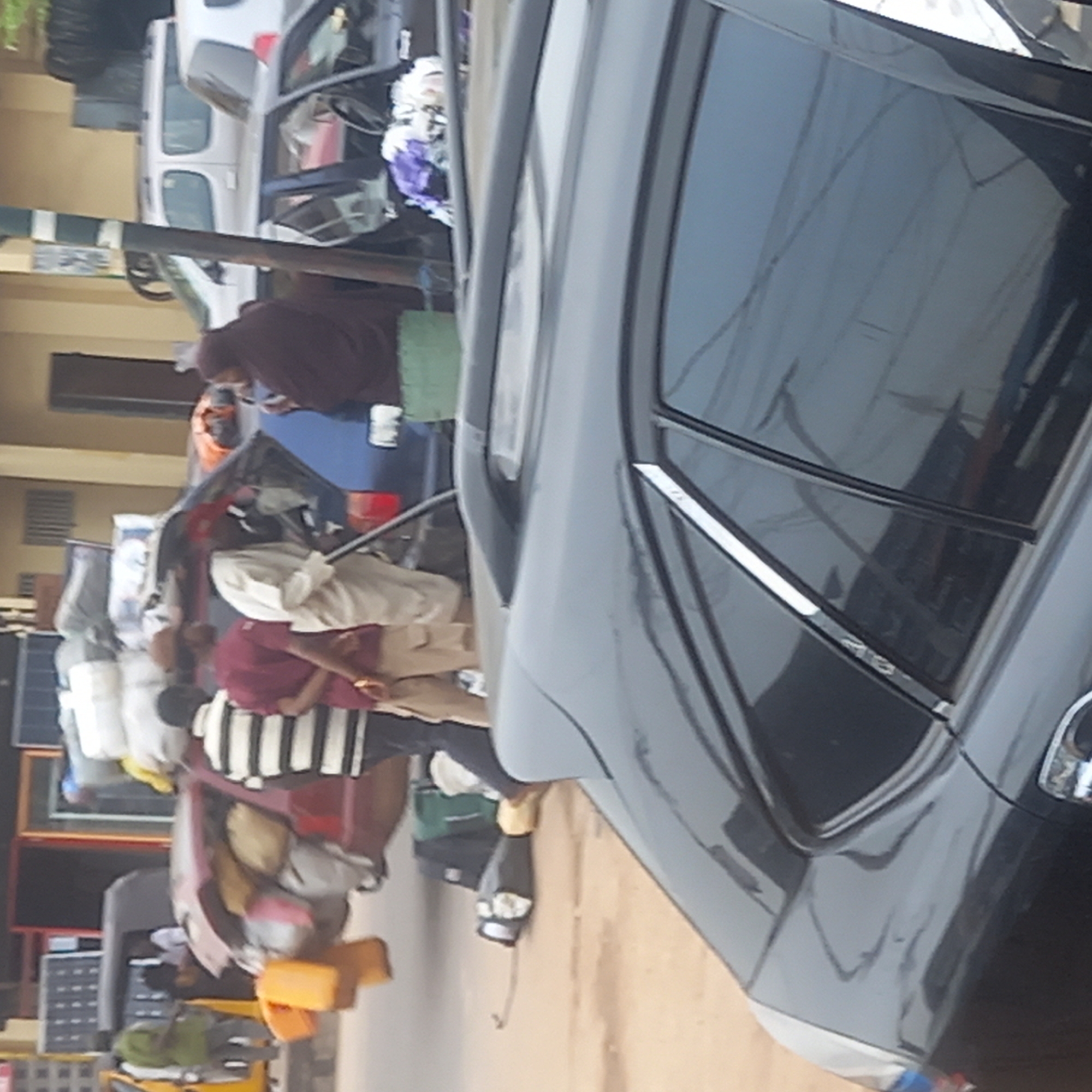 Dadi Riba, Nigeria. An urban street scene shows a congested street with multiple vehicles and pedestrians. In the foreground, the side of a dark-colored car dominates. A white van and a blue car are visible. Pedestrians are present, some carrying loads on their heads and in their hands. The scene is generally cluttered with various objects piled onto vehicles and carried by people. Buildings line the street.
