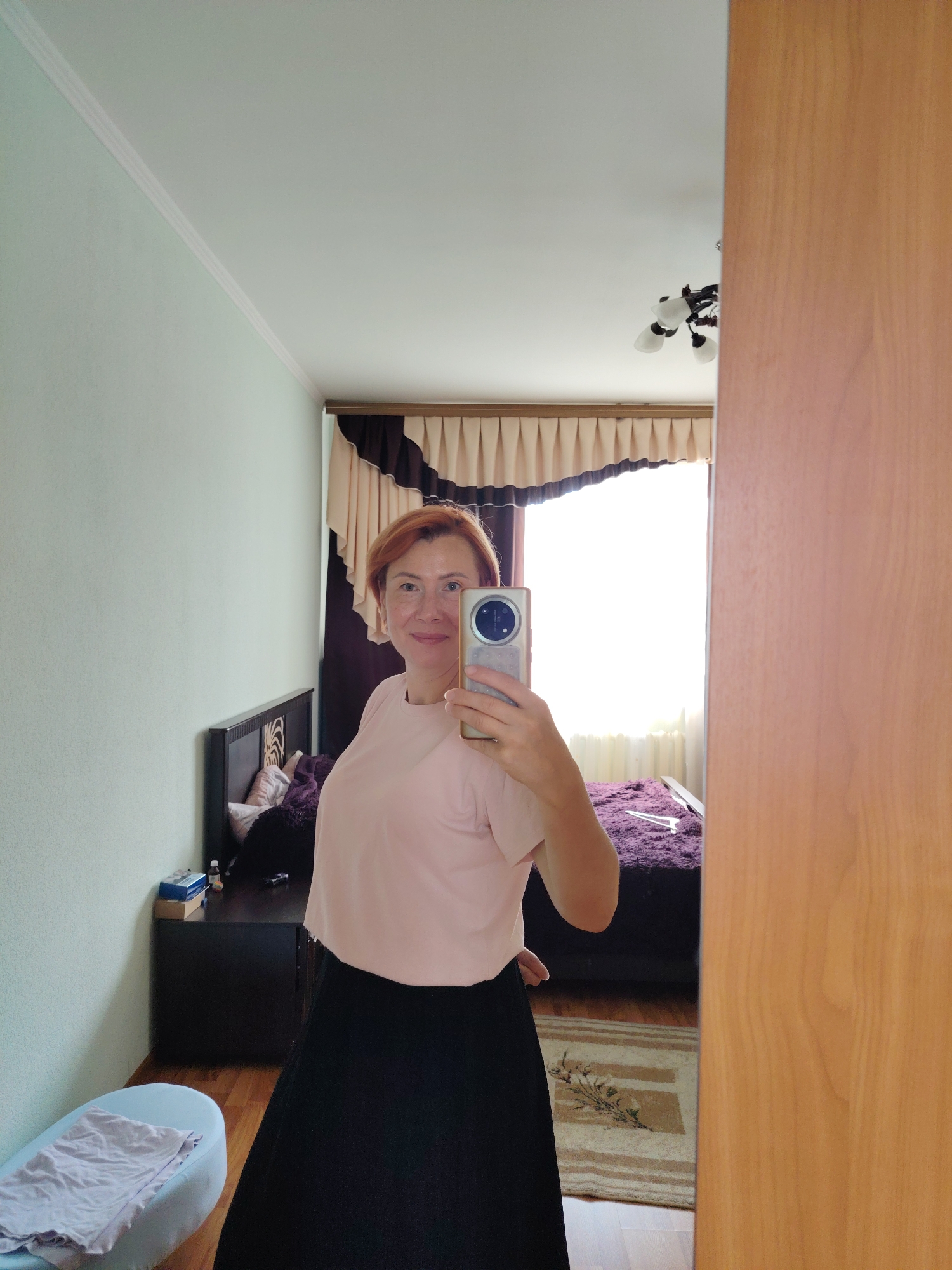 A woman with auburn hair is taking a selfie in a bedroom. She is wearing a pale pink t-shirt and a black skirt. She is holding a smartphone in her right hand. In the background, there is a bed with a purple throw, a nightstand with items on it, and a rug on the floor. A window with beige and dark brown curtains is visible behind the bed. The room has light green walls and a wooden doorframe on the right side of the image. There is also a white ceiling with a light fixture. The scene appears to be in Konakovo, Russia, given the provided context.