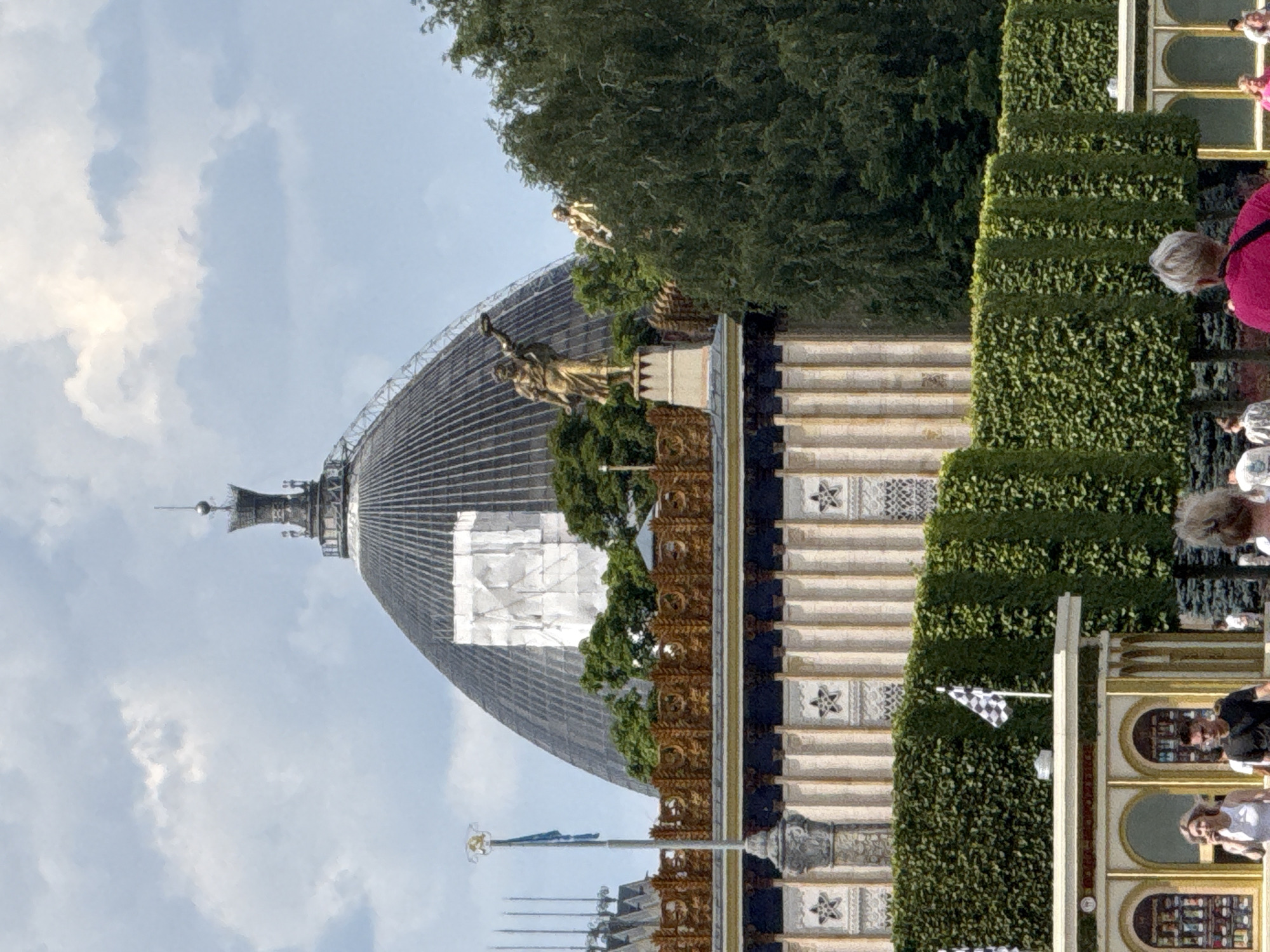 The image shows a view of the Ostankino television tower's base in Moscow, Russia, with its distinctive dome and surrounding pavilions. In the foreground, there are people walking past a series of green hedges and open-air kiosks. A checkered flag is visible on a pole behind the hedges. The background features the grand dome of the pavilion, adorned with intricate architectural details and topped with a spire. Statues are placed on pedestals, one of which is a gilded male figure in a dynamic pose. The sky is partially cloudy, with the sun casting light on the scene.