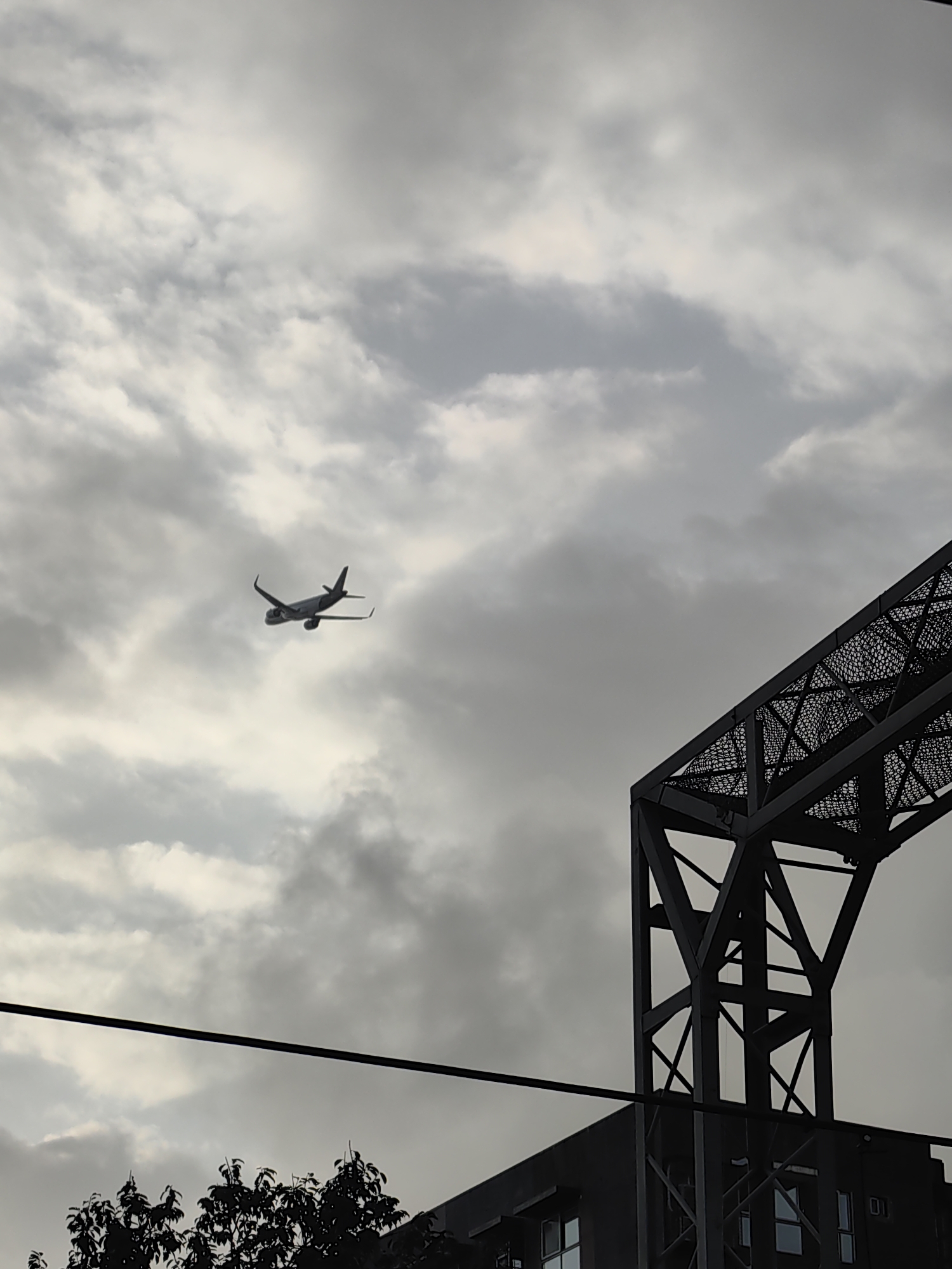 An airplane is captured in silhouette against a cloudy sky. In the foreground, a dark, geometric structure, possibly a bridge or industrial building, dominates the right side of the frame. Its metallic framework is visible, with intricate netting or lattice work. A thick black cable stretches horizontally across the lower portion of the image, with the dark silhouette of trees and a building visible below it. The scene suggests an urban environment. Given the prevalence of such infrastructure and the presence of air traffic, it is plausible that this image was taken in Mumbai, India, potentially near the Bandra-Worli Sea Link or an industrial area with a nearby airport.