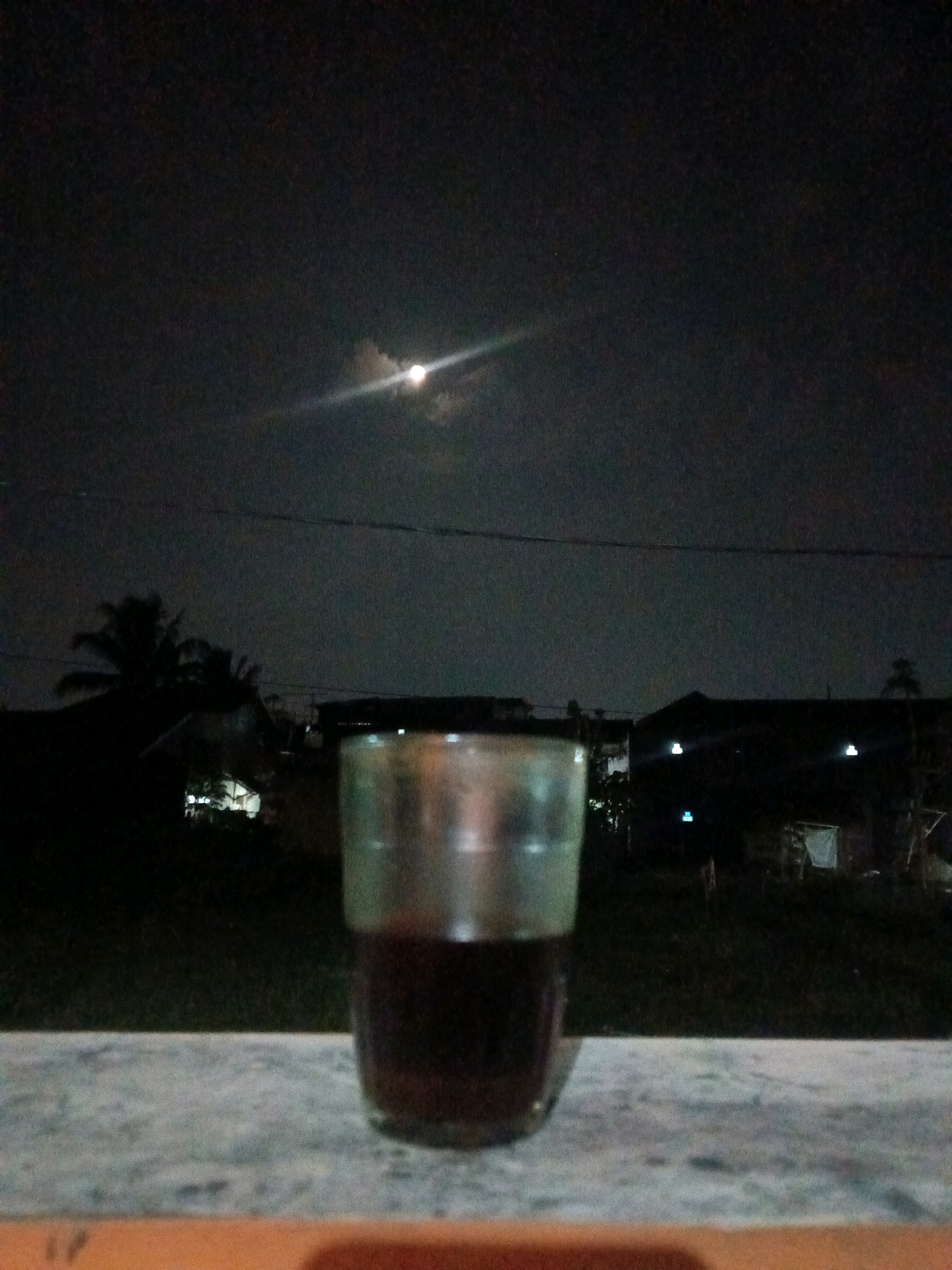 In the city of Garut, Indonesia, under a dark, cloudy night sky with a hazy moon visible, a clear glass containing a dark liquid, likely coffee or tea, rests on a textured, light-colored surface in the foreground. The glass is positioned centrally and fills a significant portion of the lower half of the frame. Behind the glass, silhouetted against the sky, are the dark shapes of trees, including a palm tree, and what appear to be low-rise buildings or houses with small, glowing lights. A thin, dark horizontal line, possibly a wire or clothesline, stretches across the upper portion of the image. The overall scene suggests a quiet evening outdoors.
