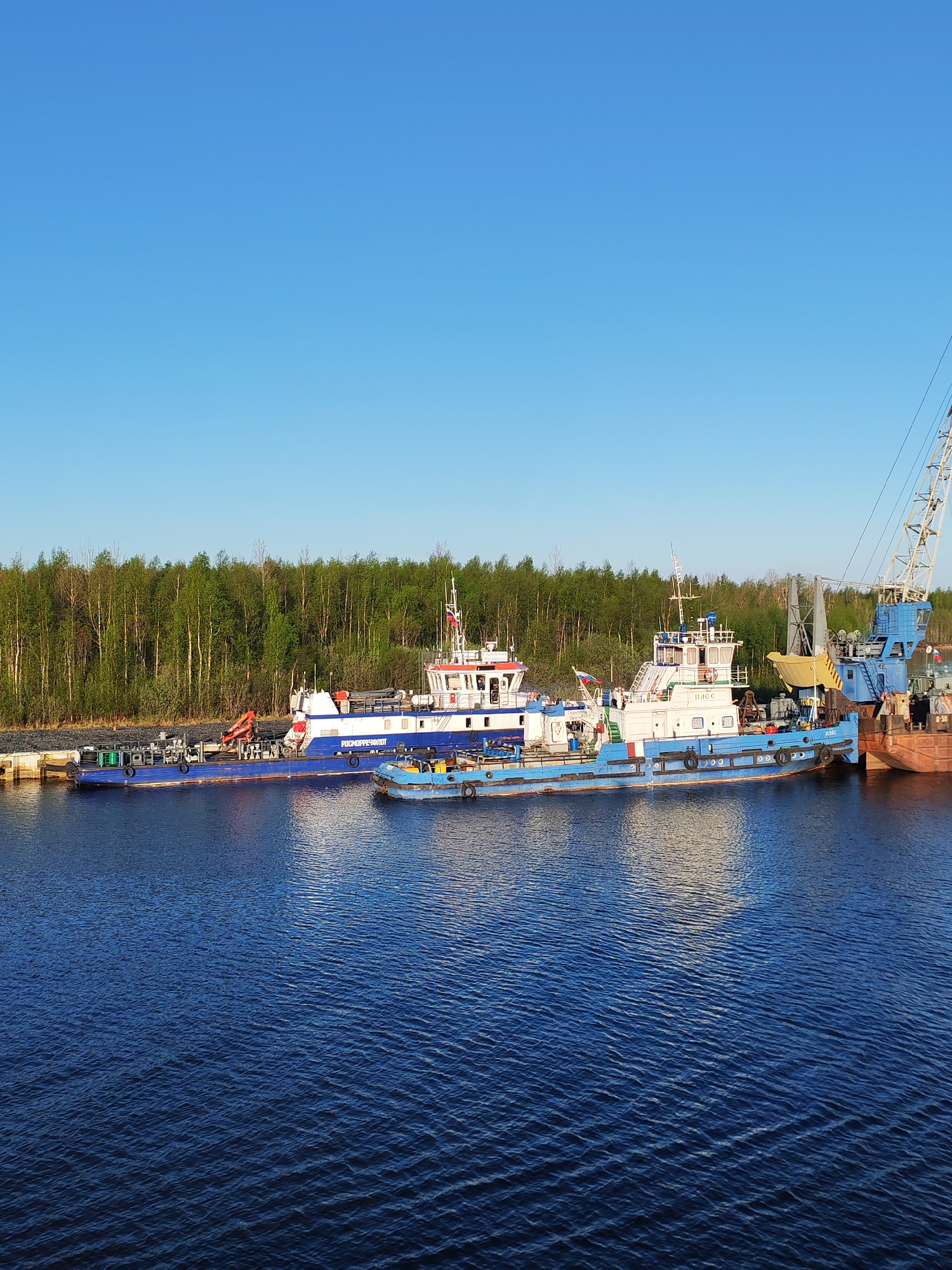 The image depicts a waterfront scene at Ozernoe Ust'e, Russia. Several boats are docked along a shoreline with a backdrop of green trees. The boats are primarily blue and white, with one showing visible Russian flags. There's a mix of vessel types, including what appears to be a crane barge or some type of industrial watercraft. The water is a dark blue, suggesting a deep or still body of water. The sky above is a clear blue, indicating a bright day. There are no visible people. The scene is static, focusing on the moored vessels and their surrounding environment.