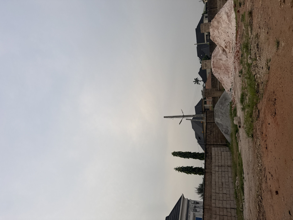 A residential area features single-family homes with dark rooftops. A pile of white sand and a pile of gray gravel suggest nearby construction. A tall power line stands prominently against the overcast sky. A brick wall borders the dirt road, along with three slender trees. The sky is overcast, creating flat lighting.
