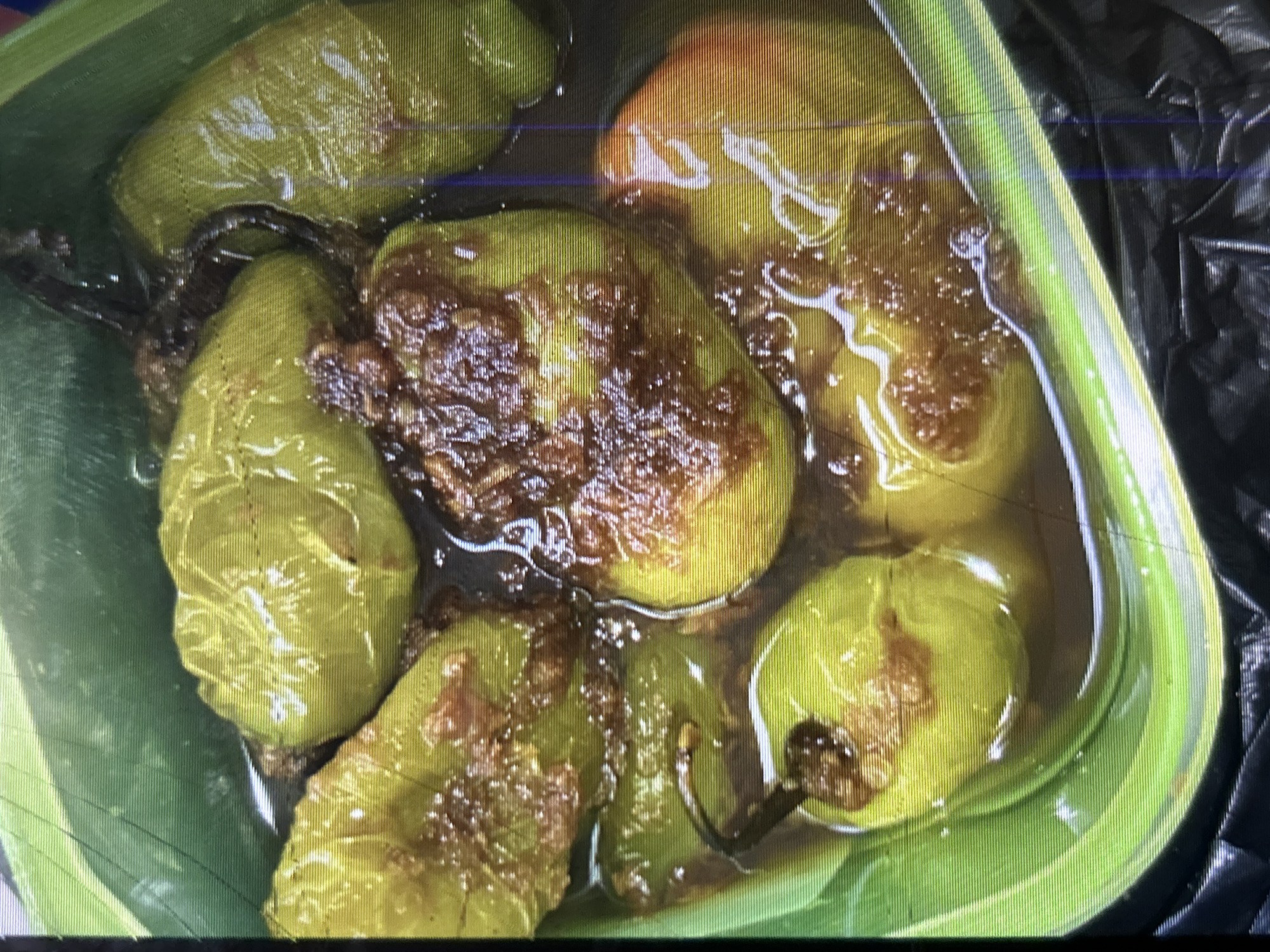 The image shows a close-up, top-down view of a green plastic container filled with several whole, cooked peppers in a dark liquid. The peppers are light green to yellowish-green in color and appear to have a slightly blistered or roasted texture. They are submerged in a savory-looking liquid, possibly a broth or sauce, with some brown, crumbly bits mixed in. One pepper on the right side of the container shows a reddish-orange hue, suggesting it might be a different variety or more cooked. The container has a ridged texture on its exterior. The background consists of a black, crinkled material, likely a trash bag. There is no indication of any specific location in Depok, Indonesia, or any people or actions taking place in the image.