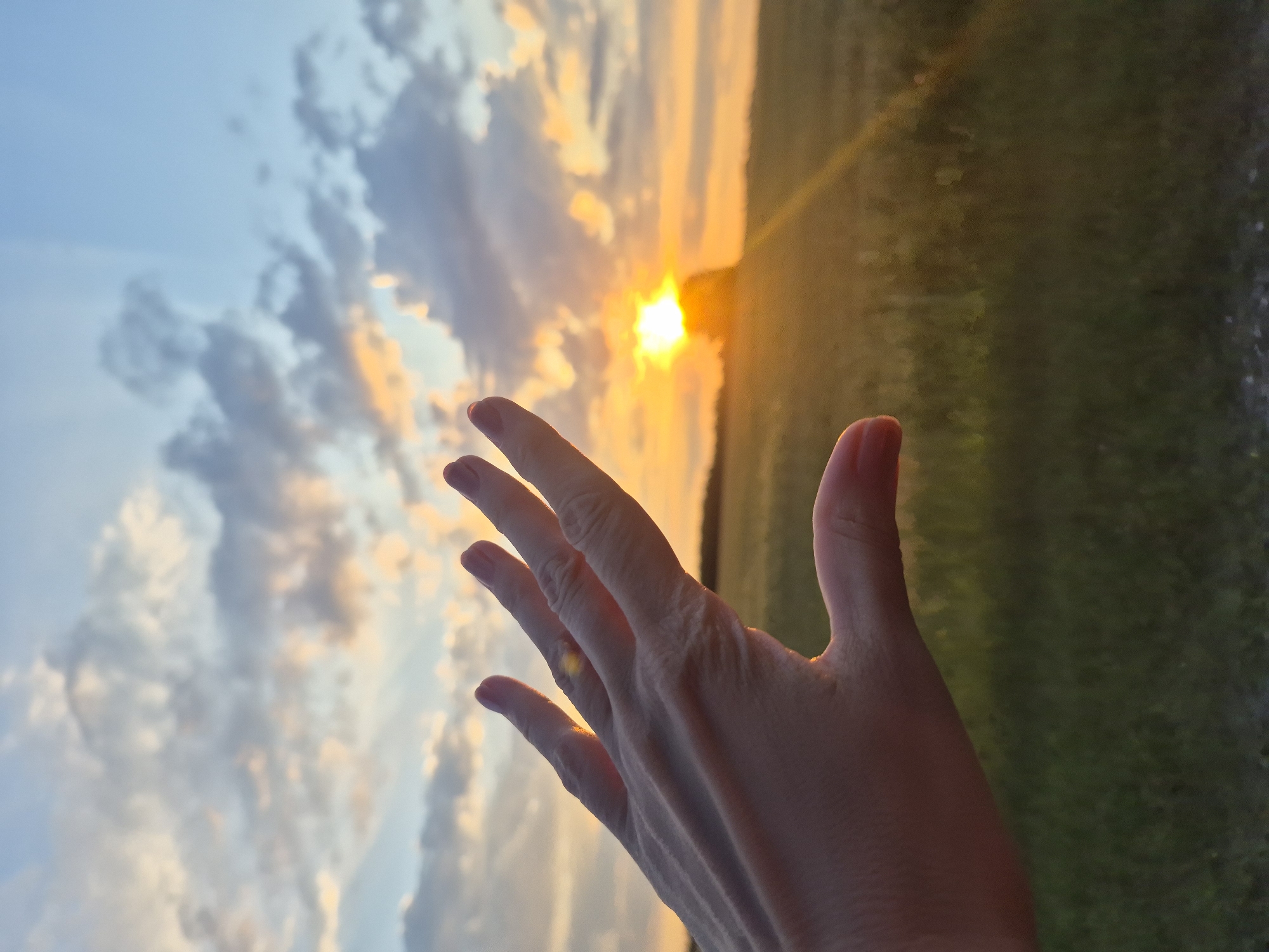 A close-up of a person's left hand is in the foreground, with fingers spread and reaching towards the right. The hand is illuminated by the warm light of a setting sun. In the background, a field is visible, bathed in the golden hues of sunset. The sky is a mix of blue and orange, with scattered clouds tinged with pink and yellow. The sun is low on the horizon, appearing as a bright, luminous orb behind a silhouetted object, possibly a tree or a distant structure, in the field. The overall impression is one of serenity and the beauty of a natural landscape at dusk. The image is identified as being located in the city of Ol'khovo, Russia.