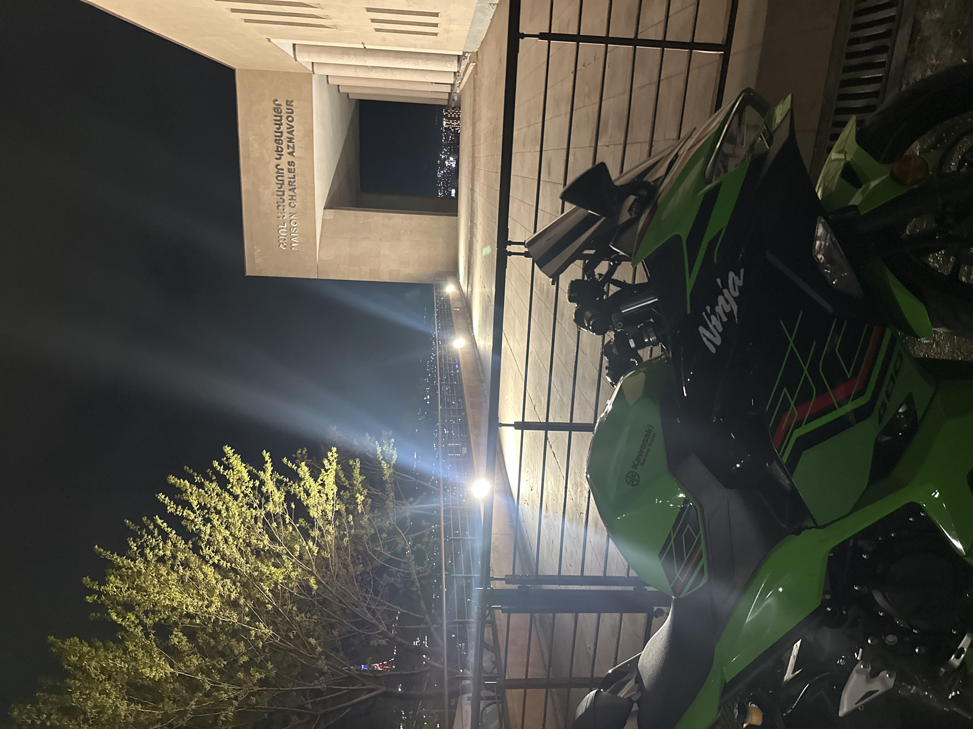At night, a green and black Kawasaki Ninja motorcycle is parked in Yerevan, Armenia, with the Maison Charles Aznavour building in the background. The building, featuring the name "Charles Aznavour" in both Armenian and English, is illuminated. A tree with glowing foliage is visible to the left. Fencing is also visible between the motorcycle and the Maison Charles Aznavour building.