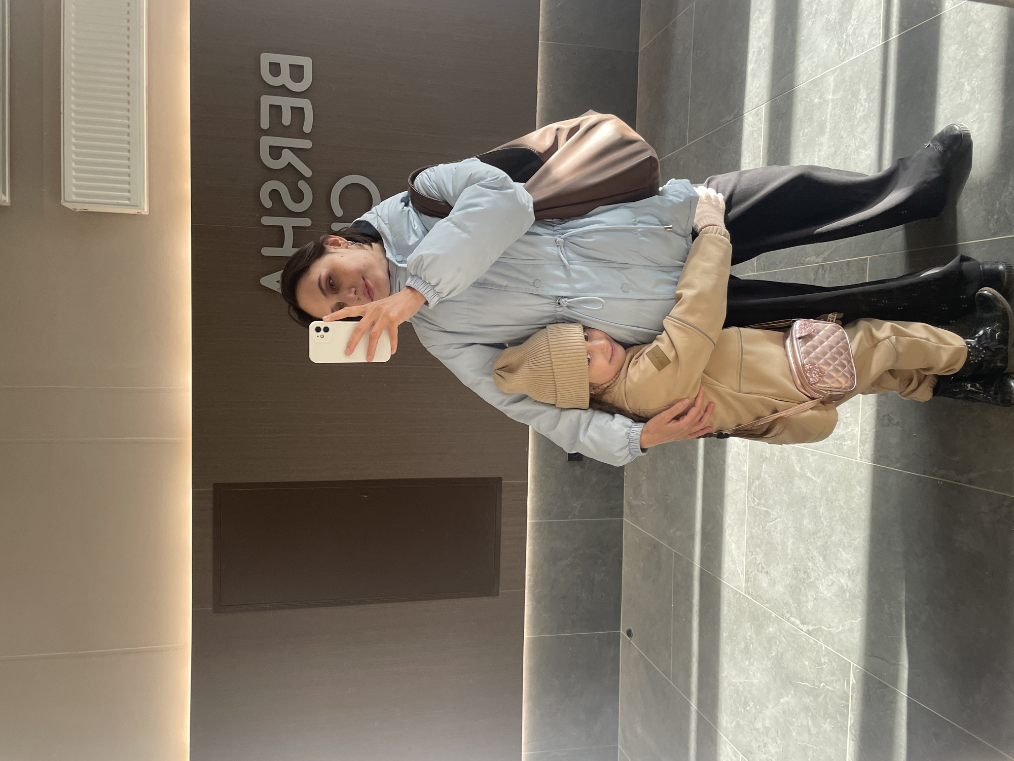 An adult woman and a child are posing for a selfie indoors, likely within a commercial establishment. The image is rotated 90 degrees clockwise. The woman, on the left, has dark hair and wears a light blue puffer jacket, dark pants, and dark boots, with a brown shoulder bag slung over her. She holds a white smartphone, extending her arm to take the photo, and has a slight smile. The child, a young girl, stands to the woman's right, wearing a beige puffer jacket, a matching knit beanie, beige pants, and black boots. She carries a small, light pink quilted crossbody bag and is hugging the woman's waist, also smiling at the camera.
The background features a dark brown paneled wall with large white letters that, when corrected for rotation and reflection, spell "BERSHKA". This suggests the photo was taken in a store of the fashion retailer Bershka. Adjacent to this, a lighter beige wall has a white rectangular ventilation grate or radiator, and a vertical strip of warm light illuminates the space between the walls. The floor is composed of large dark grey tiles, displaying distinct patterns of sunlight and shadow, indicating it is daytime. The overall scene captures a joyful moment between the two individuals in a Bershka store located in Izhevsk, Russia.