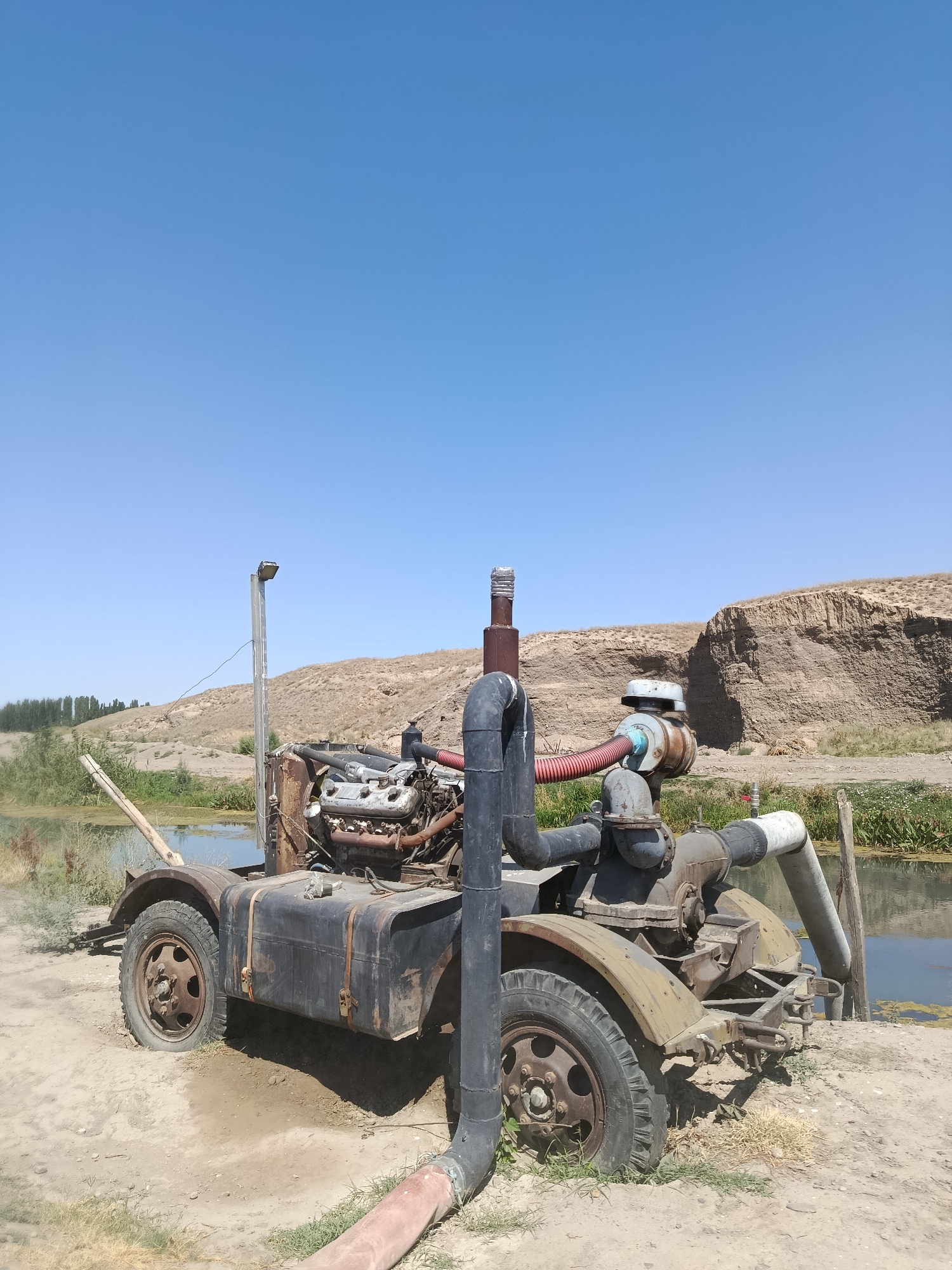 In the city of Kenes, Kazakhstan, an abandoned water pump sits on the muddy bank of a body of water. The pump, mounted on a wheeled trailer, appears to be powered by an old engine. Large pipes extend from the pump, one feeding into the water and another exiting it. The scene is desolate, with dry, cracked earth and sparse vegetation surrounding the water. In the background, a large, eroded cliff face looms under a clear blue sky. The overall impression is one of decay and disuse, with nature slowly reclaiming the man-made structure.