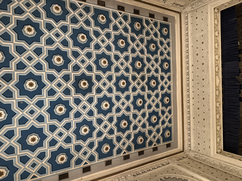 The image depicts the ornate ceiling and upper wall of the Mirror Hall within the National Opera of Ukraine (Kyiv Opera House), located at Volodymyrska Street 50, Kyiv, Ukraine. The ceiling exhibits a complex geometric tessellation composed of interlocking eight-pointed stars and square/octagonal shapes, rendered primarily in deep blue and light gray. The center of each prominent eight-pointed star contains a smaller, stylized, multi-layered circular motif with a golden-brown core. Below the ceiling, the upper wall and cornice feature elaborate relief stucco work in an off-white or cream color, showcasing intricate arabesque patterns, scrolled motifs, a horizontal band with a repeating pattern of small, raised dots, and decorative shell-like carvings along a lower molding. A dark blue, pleated curtain, consistent with stage drapery or heavy window coverings, is partially visible at the bottom of the frame. No individuals are present.