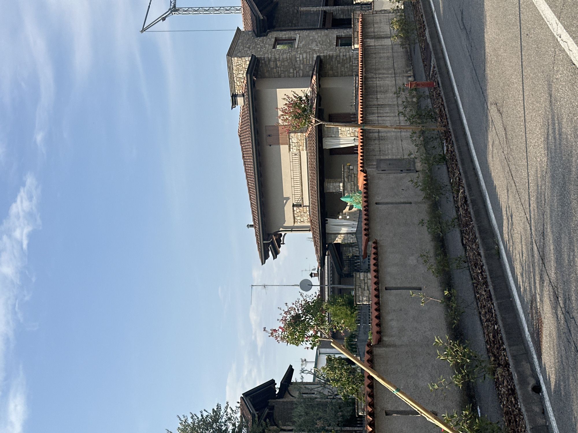 In the city of Rudiano, Italy, this image captures a residential street scene under a clear blue sky with scattered white clouds. A grey concrete wall with a red-tiled sawtooth-patterned coping runs horizontally across the foreground, separating the road from private properties. Several small, green-leafed shrubs are planted in a mulched strip between the wall and the asphalt road. A bright red fire hydrant is visible on the right side of the image.
Behind the wall, several modern houses are visible. The central house features a beige facade with stone accents and a red-tiled roof. It has a covered porch with white curtains and a second-story balcony with a black metal railing. A thin tree with pinkish-red blossoms stands in front of this house. To the left, another house with a darker stone facade and a steeply pitched roof is partially visible. A tall, thin wooden pole leans diagonally across the left side of the frame, positioned in front of the wall and the shrubs. In the background, a construction crane is visible against the sky, suggesting ongoing development. The road in the foreground is asphalt, marked with a white line.
