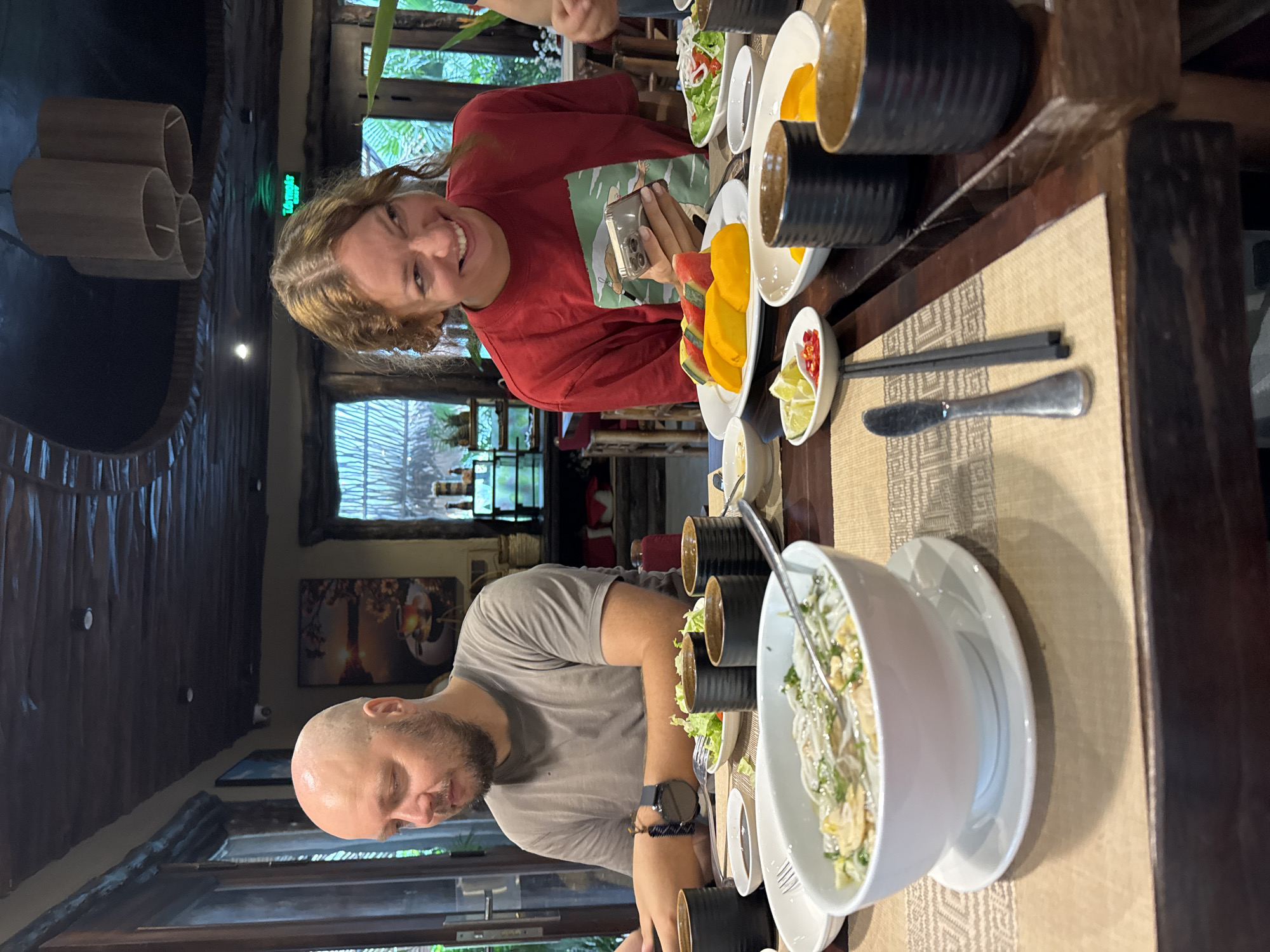 A man and a woman are seated at a wooden table in a restaurant. The man, with a shaved head and beard, is wearing a grey t-shirt and looking down at a watch on his left wrist. The woman, with blonde curly hair, is wearing a red t-shirt and smiling while looking at a smartphone in her hands. In front of them are bowls of food, including a bowl of soup with noodles and vegetables, a plate of fruit with mango and watermelon slices, and small dishes with limes and chili peppers. Several black ceramic cups are also on the table. The restaurant appears to have a rustic ambiance with wooden paneling on the ceiling and walls, and decorative lamps hanging from above. The setting suggests a meal being enjoyed in a relaxed atmosphere, possibly in a tropical location given the fruit.