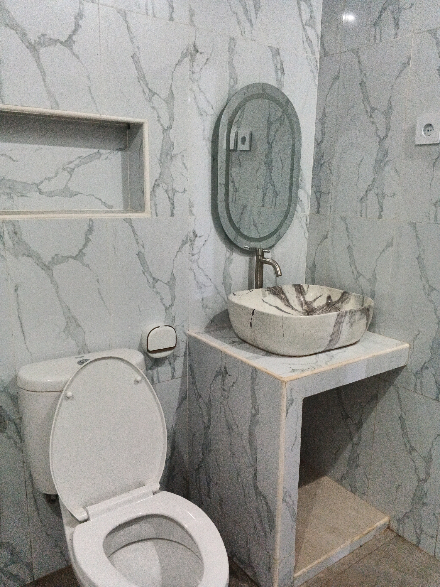 The image depicts a bathroom with marble-patterned walls and a countertop. On the left, a white toilet with an open lid is visible. To the right of the toilet, a toilet paper holder is mounted on the wall. A sink with a marble pattern sits on the countertop, accompanied by a silver faucet. Above the sink, an oval mirror with an illuminated frame is hung on the wall. An electrical outlet is present on the upper right wall. A recessed shelf is built into the wall above the toilet. The floor is a neutral gray color. The scene is a photograph of a bathroom. The location is Sirau, Indonesia.