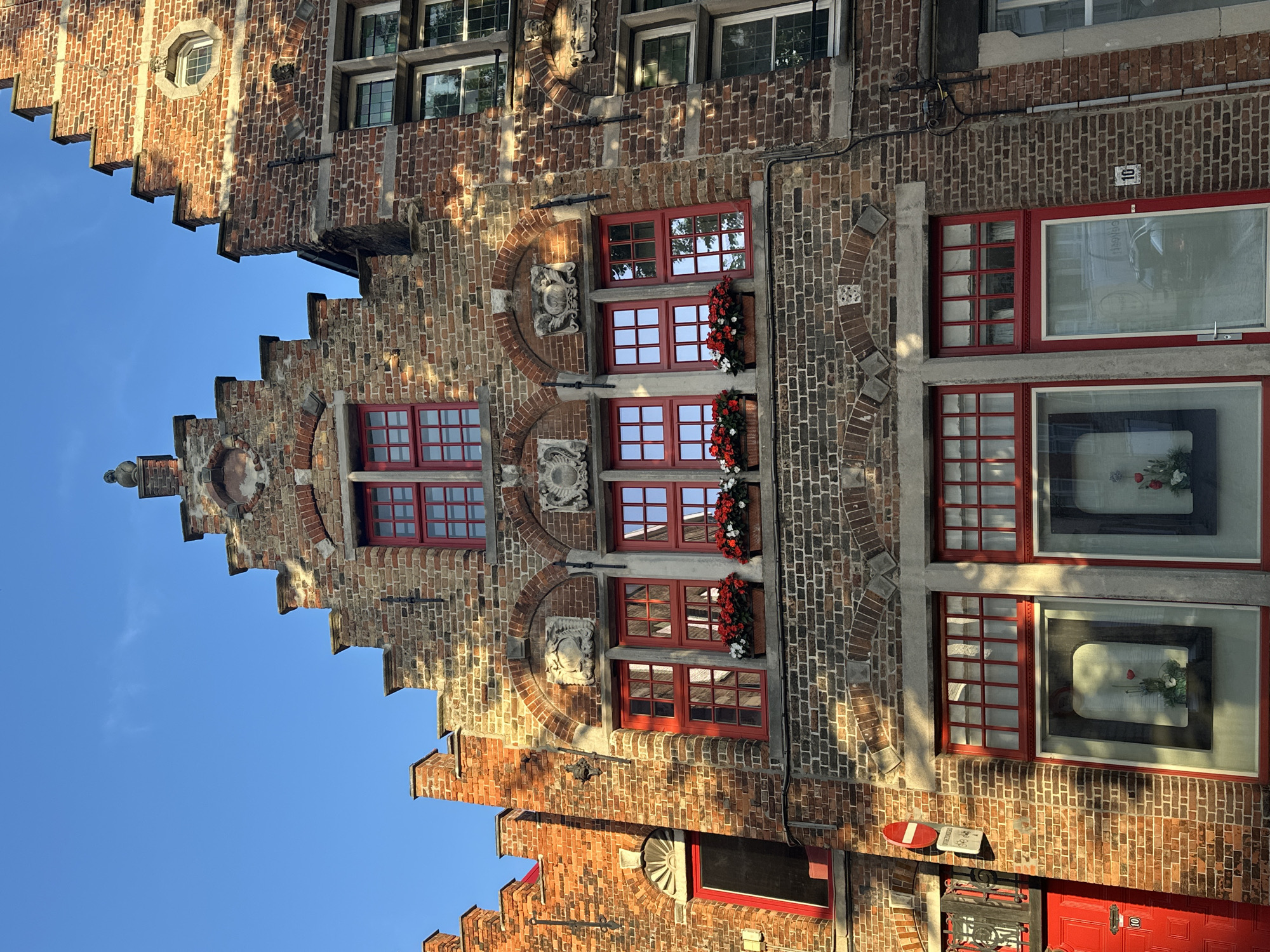 The image captures a close-up view of the facades of historic brick buildings in Bruges, Belgium. The architecture features characteristic stepped gables, red-framed windows with multiple panes, and decorative stone carvings above some windows. Red flowers in window boxes add a touch of color to the brickwork. The buildings appear to be at street level, with shop windows visible on the lower floors. One window displays a floral arrangement. Red doors and signage are also present. The bright sunlight suggests it is daytime, and the clear blue sky indicates fair weather. The overall impression is one of a charming, well-preserved historic street in Bruges.