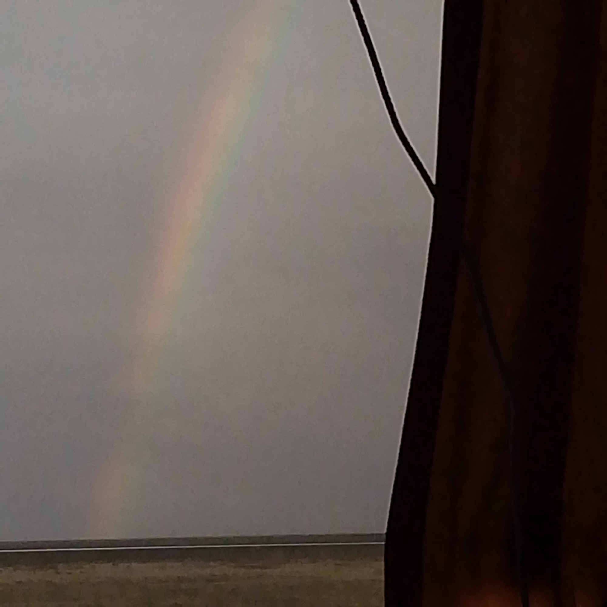 The image depicts a faint rainbow arcing across a grey, overcast sky. The rainbow is visible in the upper left quadrant of the frame. In the foreground, the right side of the image is dominated by a dark, vertical, textured object that appears to be a wooden post or structure. A thin black line, possibly a wire, runs diagonally from the top right towards the center of the image, near the rainbow. Below the sky, a horizontal line marks the horizon, with a muted, brownish landscape beneath it, suggesting a rural or undeveloped area. The overall impression is of a natural phenomenon observed under a somber sky, possibly from a sheltered vantage point. No elements in the image suggest a specific location within Sarul, Russia.
