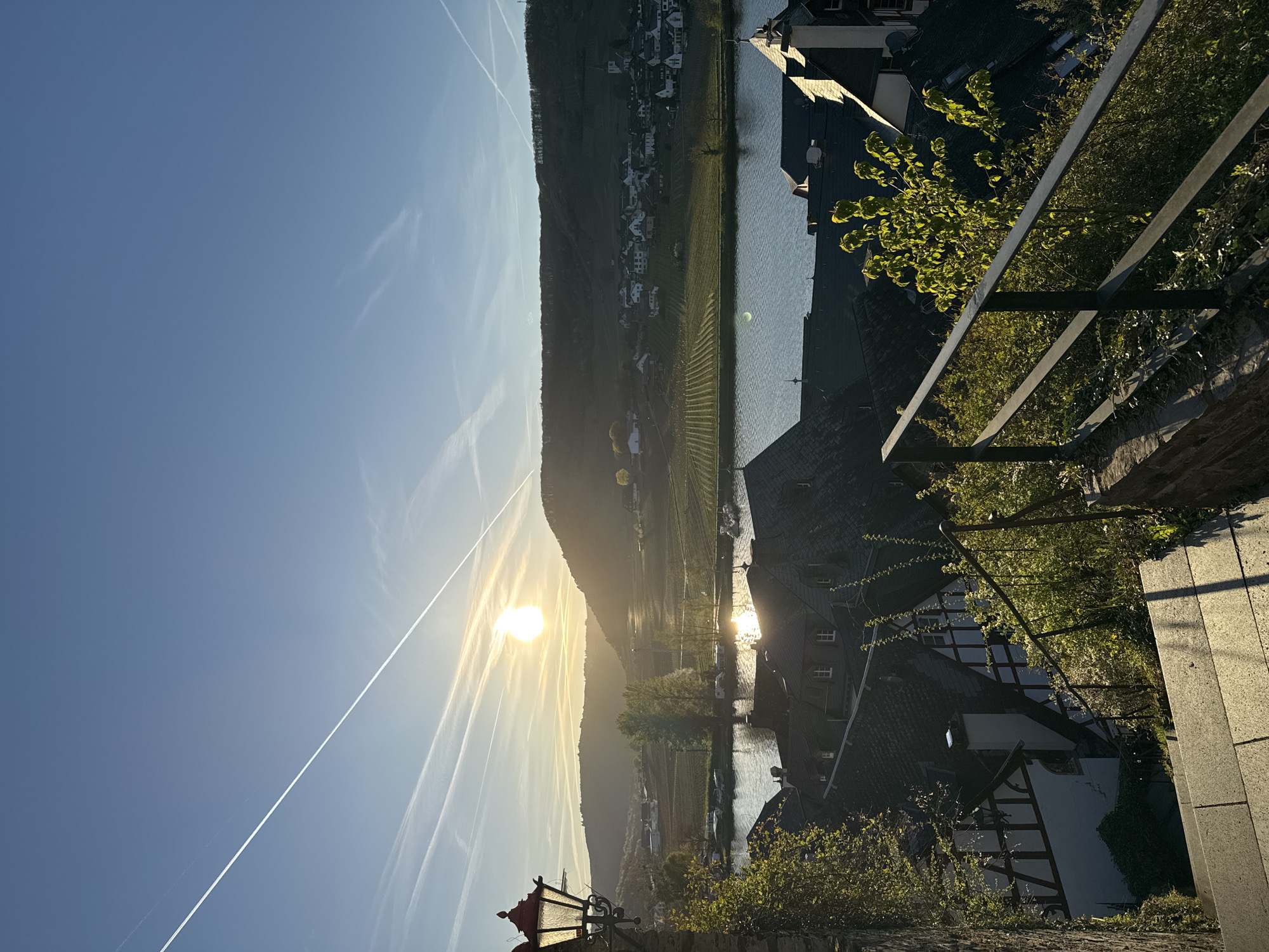 Here is a description of the image:
The image shows a view in Beilstein, Germany, with a hill covered in vineyards on the opposite side of a river. The sun is visible near the center of the image, shining over the hill and reflecting on the river. Contrails streak across the blue sky. In the foreground, there is a stone wall and a metal railing, along with some greenery. Several traditional-looking buildings with dark roofs and light-colored walls are visible closer to the river. A portion of a red lamppost is visible in the lower left corner.