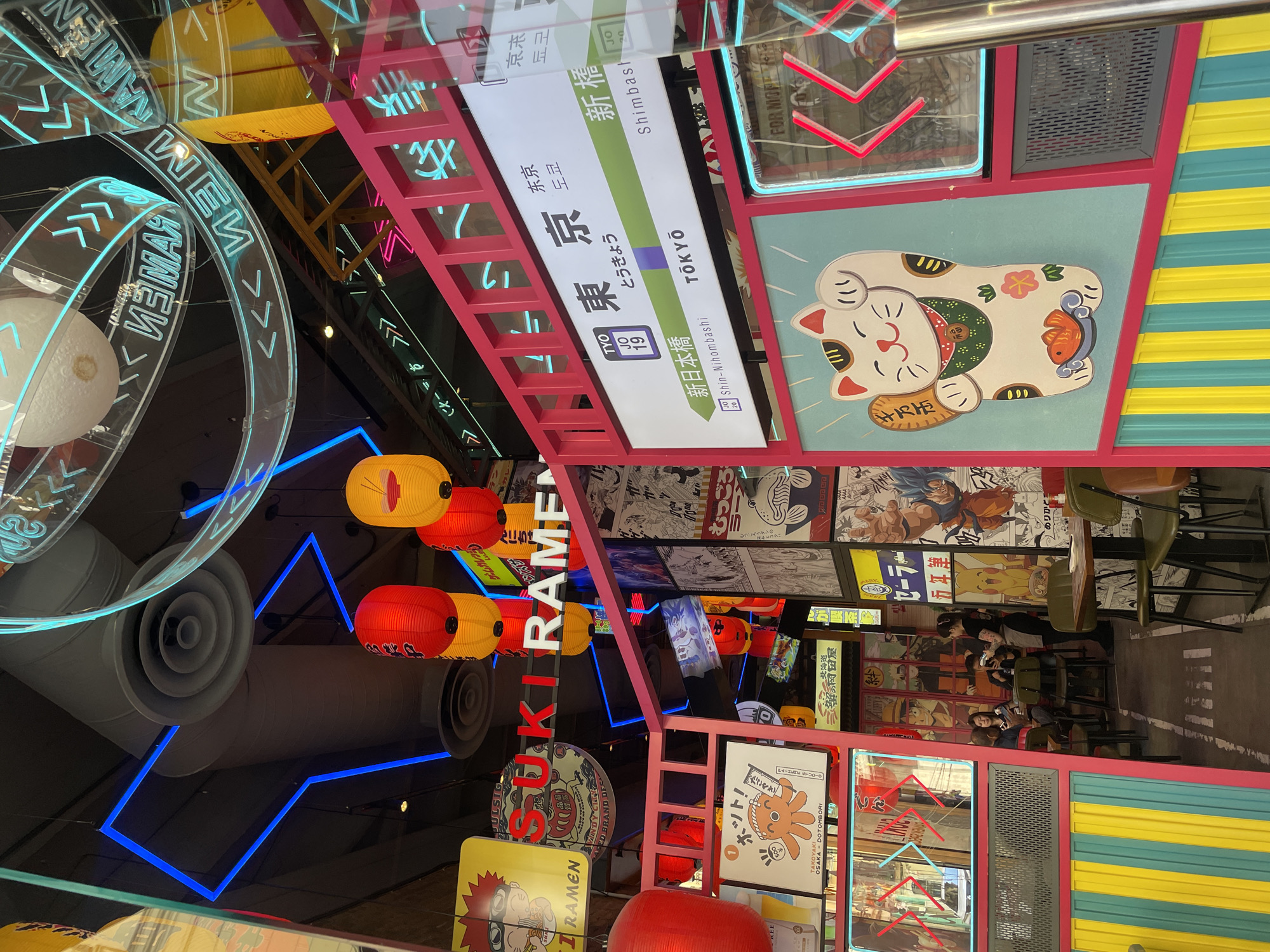 The image is an interior shot of a restaurant. The ceiling is decorated with lights, lanterns, and signage, some of which read "SUKI RAMEN." There is a faux Japanese train station sign that includes the names "Tokyo," "Shin-Nihombashi," and "Shimbashi." A maneki-neko (beckoning cat) decoration is visible. The walls are adorned with manga and anime panels. There are tables and chairs, and several people can be seen seated at one of the tables. The restaurant is located in Barcelona, Spain.