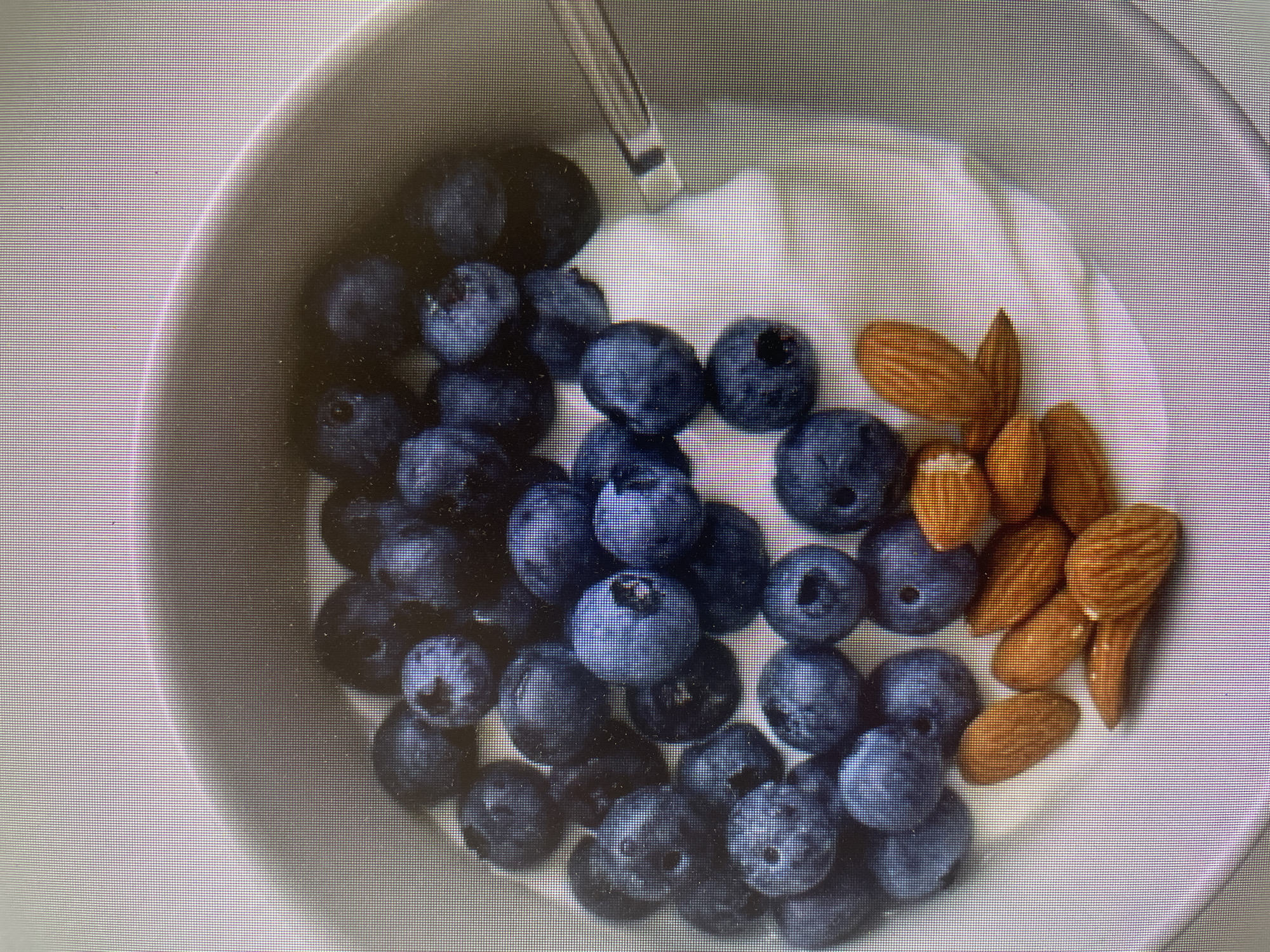 A top-down shot captures a white bowl filled with a healthy snack or meal. The bowl contains a generous portion of white yogurt, with an assortment of blueberries and almonds arranged on top. A pile of dark blue blueberries dominates the left and central parts of the bowl, while a cluster of light brown almonds occupies the right side of the yogurt. A silver spoon handle is partially visible, extending from the top center towards the yogurt. The lighting is bright and even, suggesting an indoor setting. There are no people visible, nor are there any discernible location cues specific to Poltava, Ukraine, time of day, weather, or visible text. The image itself exhibits a screen-door effect, indicative of a photograph taken of a digital display.