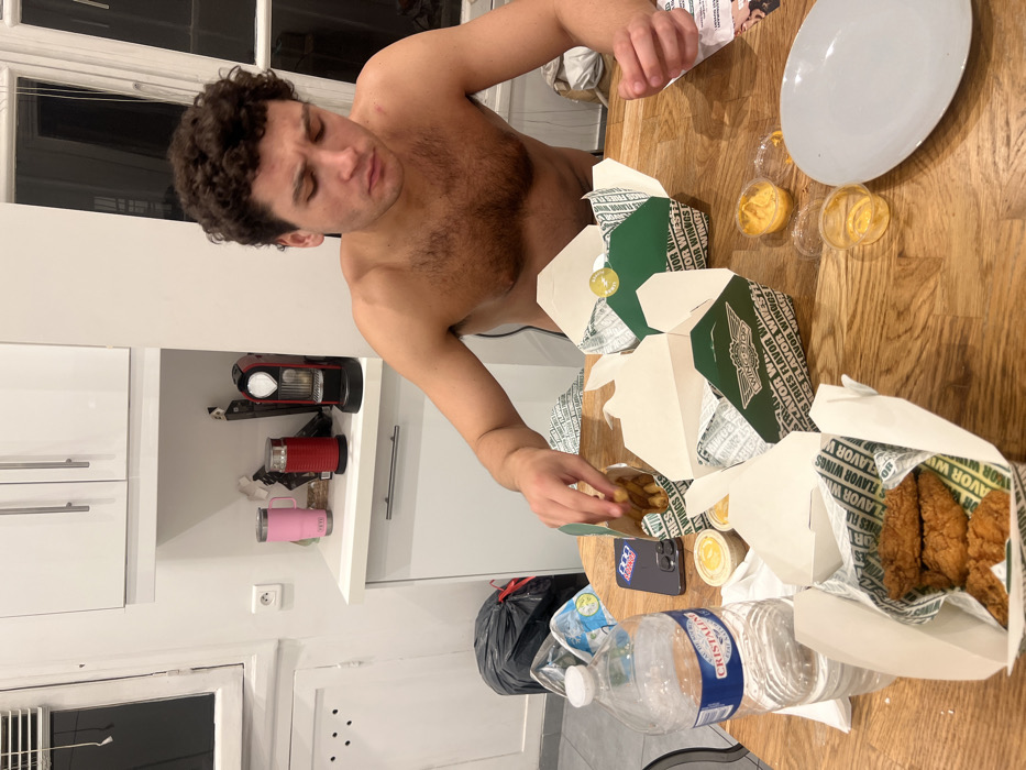 A shirtless man sits at a table covered with Wingstop takeout boxes, sauces, a plate, a phone, and water bottles. The boxes are green and white, with the Wingstop logo. The food inside includes chicken tenders and fries. The man is reaching for the fries. A white cabinet with appliances on a shelf sits in the background. A trash bag is on the floor.