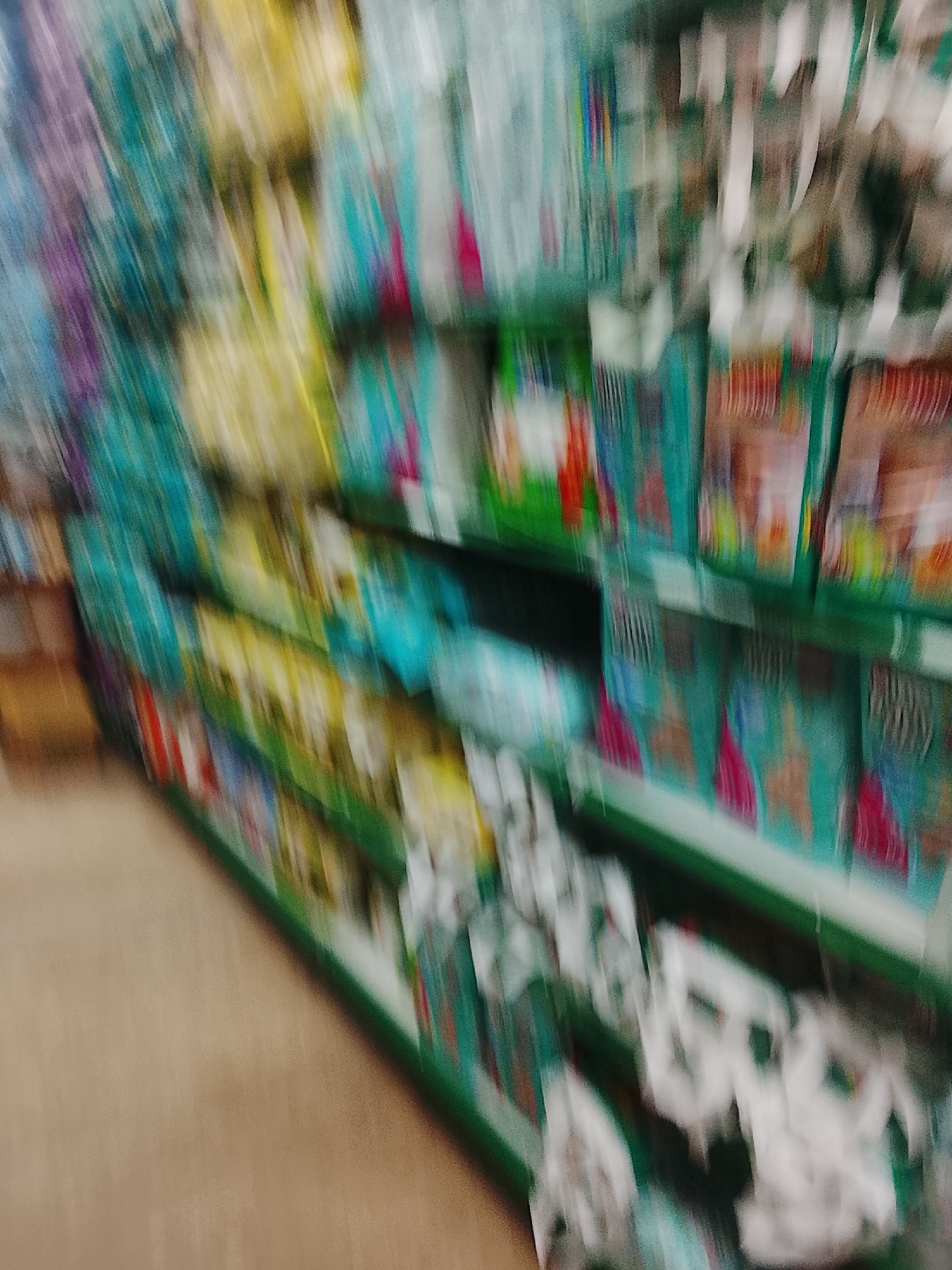 The image depicts a blurred view of supermarket shelves stocked with various products. The products on the shelves include items in teal packaging with red, green, and brown accents, along with yellow packaged goods. At the bottom right corner, white items are stacked. Based on the location provided, it is likely a retail store in Gulgasht Colony, Pakistan. Due to the blurring, specific items and brand names are indistinguishable. No individuals are visible in the scene.