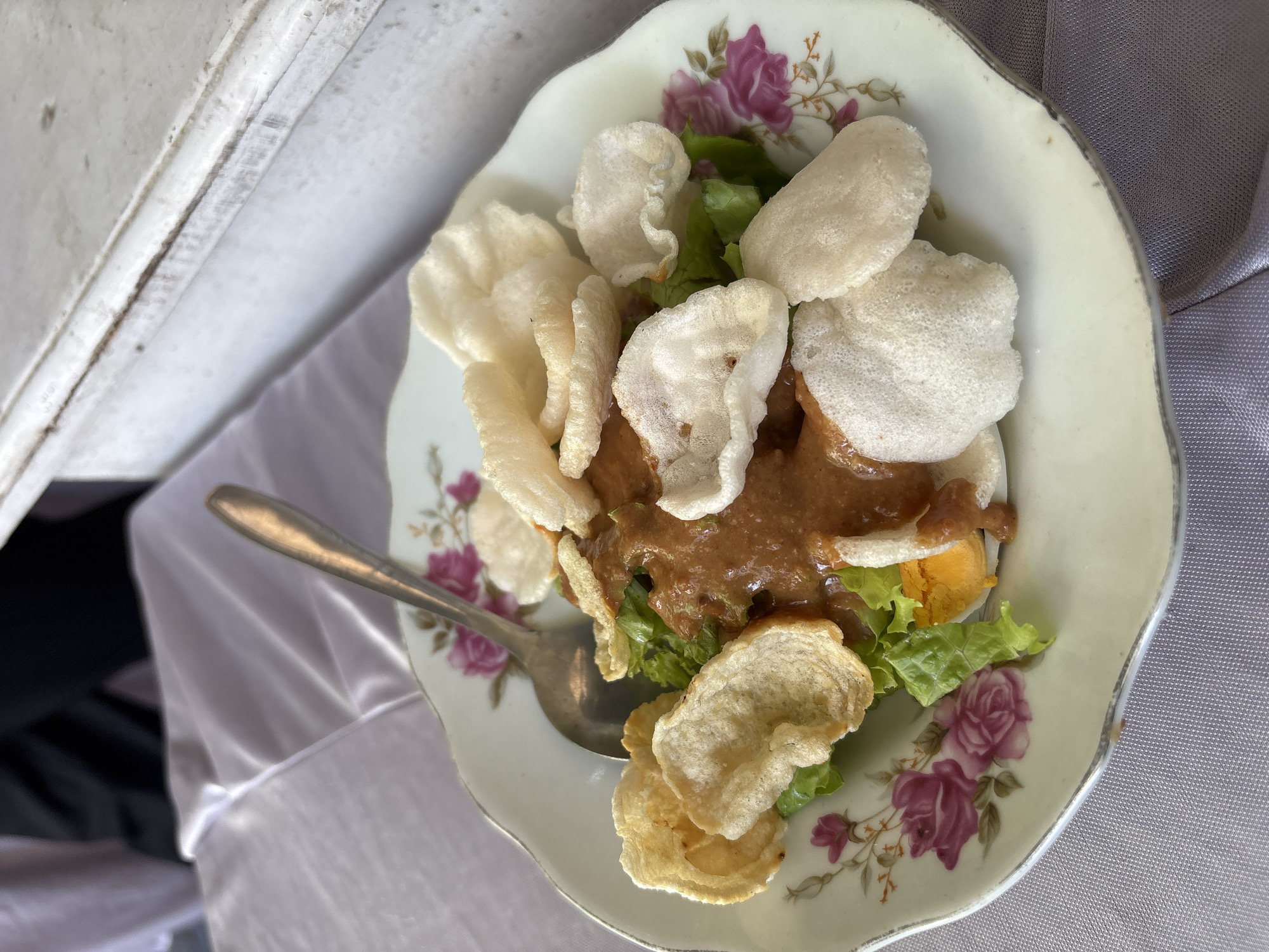 A high-angle, close-up photograph captures a single serving of Gado-gado, a traditional Indonesian salad, presented on a decorative plate in Wonocolo, Indonesia. The plate, light cream in color with a scalloped edge, is adorned with a floral pattern of pink roses and green foliage. Centered on the plate, a bed of vibrant green lettuce leaves is visible beneath a generous application of thick, reddish-brown peanut sauce. Portions of hard-boiled egg are discernible, partially covered by the sauce. The dish is topped with an assortment of Indonesian crackers (krupuk); some are light beige, irregularly puffed, while others are flatter and off-white. A silver-colored metal spoon rests on the left side of the plate, partially obscured by crackers. The plate is positioned on a light lavender-grey fabric surface, likely a tablecloth, exhibiting a subtle sheen and soft folds. The background is softly blurred, showing a dark, indistinct form in the upper left and a vertical, light-colored, textured surface on the right.