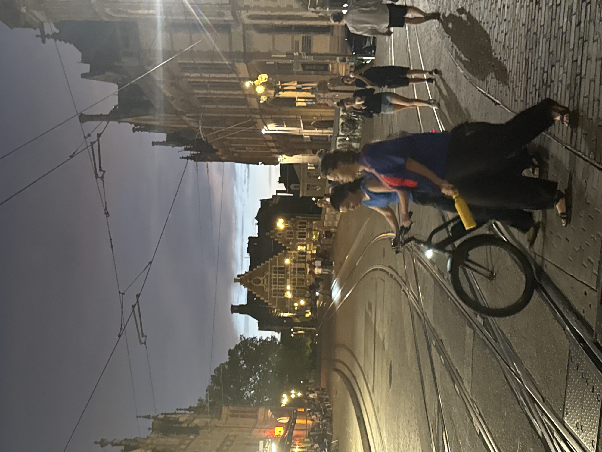 The image depicts a street scene in Ghent, Belgium, at dusk. The foreground features a bicycle with a yellow object attached to its frame, being ridden on tram tracks. Two women are in the process of crossing the street, with one pushing the bicycle. In the background, the historic architecture of Ghent is visible, illuminated by streetlights and the fading twilight. Several other individuals are walking on the cobblestone sidewalk. The sky is a deep blue, indicating the transition from day to night, and overhead wires for the tram system are prominent. The scene captures a typical evening in the city, with a blend of everyday activities and historical ambiance.