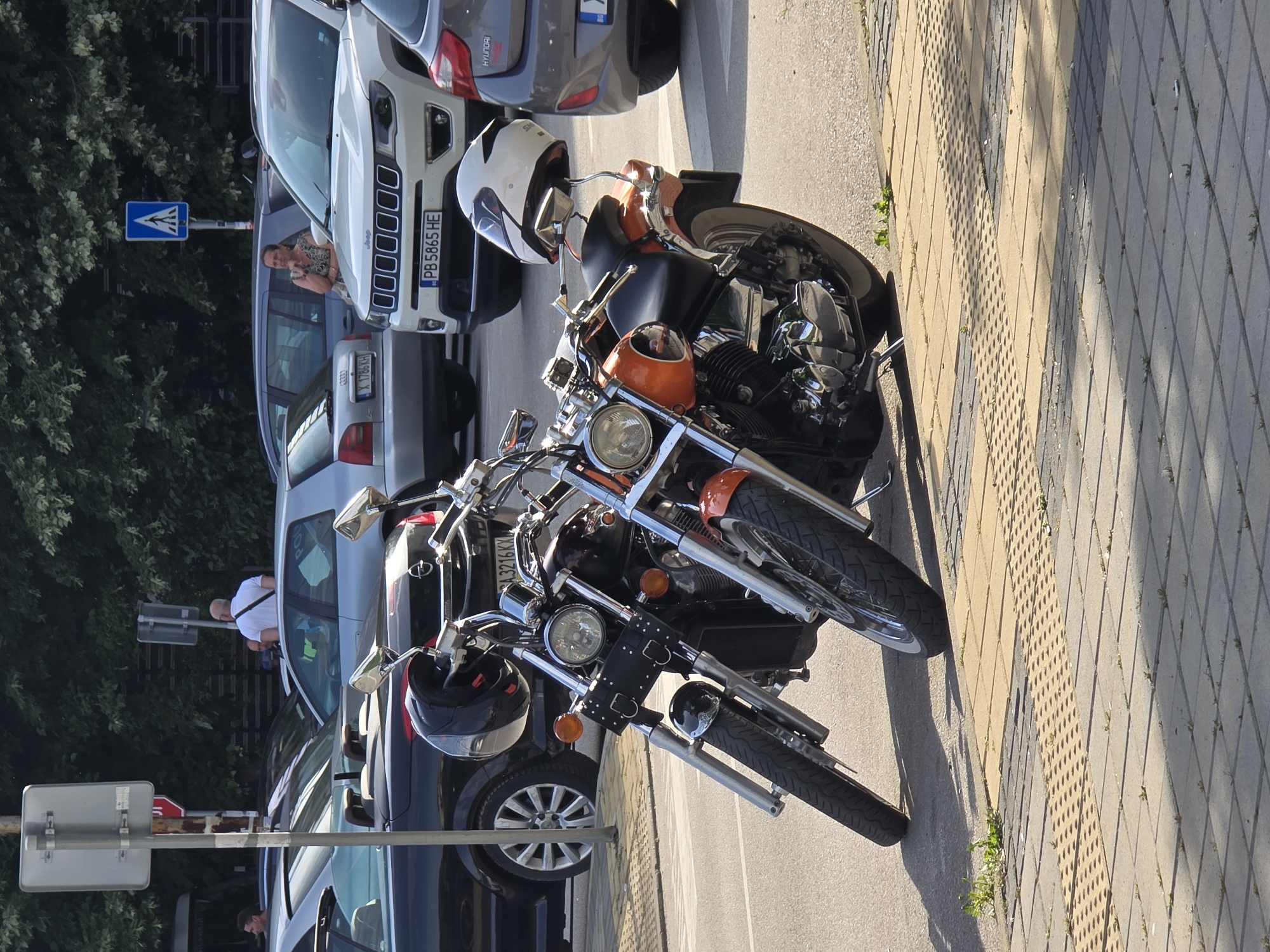 Here's a description of the image:
The image shows two motorcycles parked on a paved area near a street in an urban setting. The motorcycles are positioned close together. The first motorcycle has an orange fuel tank and a white helmet resting on the handlebars. The second motorcycle has a black helmet on the handlebars. The registration plates on the cars parked behind the motorcycles are Bulgarian. One person is visible sitting in one of the cars, and another is standing in front of another car. Street signs are also visible in the background.