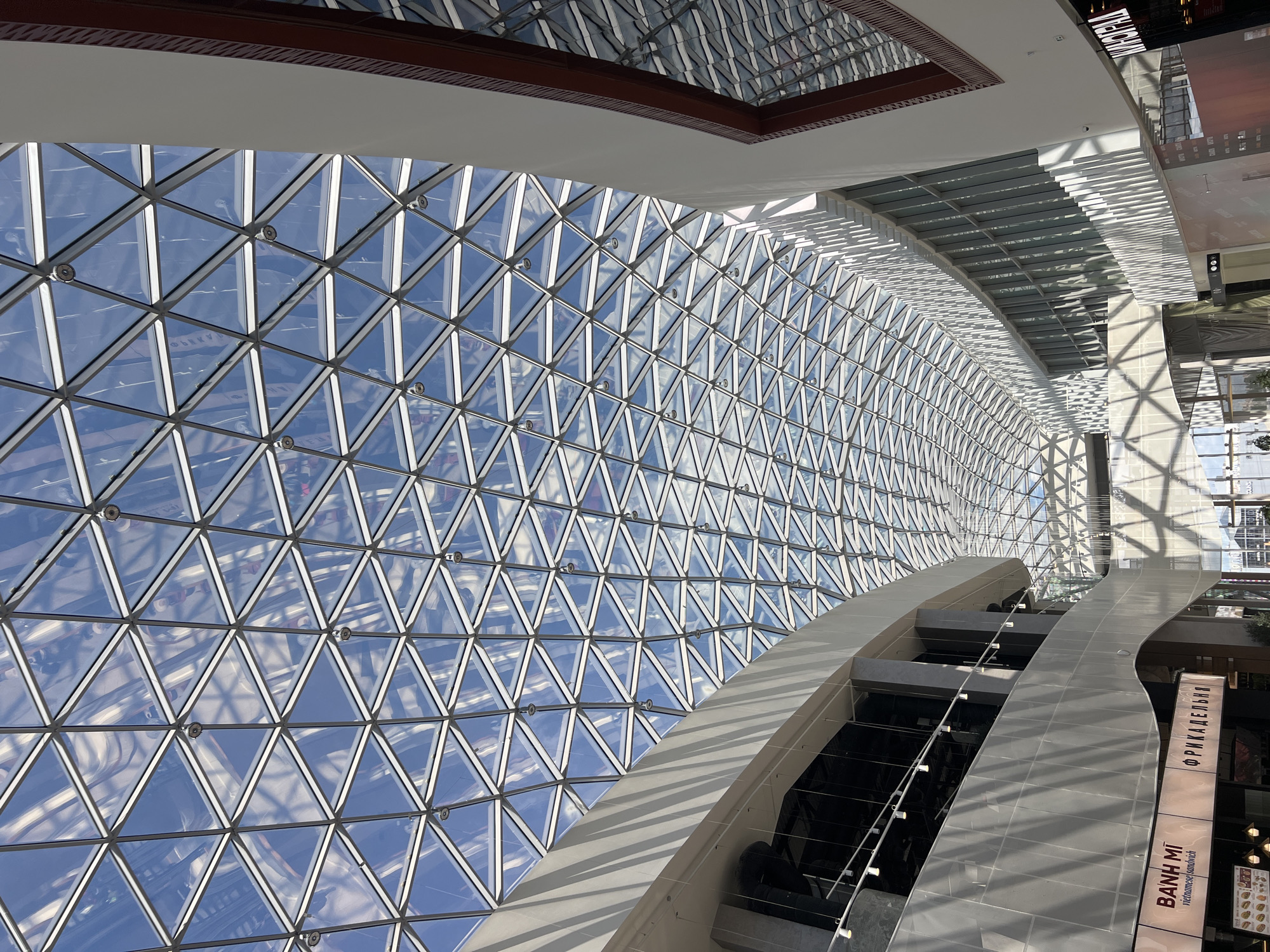 The image shows the interior of a modern shopping mall in Saint Petersburg, Russia, with a distinctive geodesic glass and metal dome ceiling. The ceiling is a complex geometric structure composed of numerous triangular panes of glass supported by a network of white metal struts. Sunlight streams through the glass, casting intricate patterns of light and shadow onto the mall's interior.
On the left side of the frame, a curved, multi-story structure with a white facade dominates the lower portion. This structure appears to house various shops and businesses, with signage visible at ground level, including "BANH MI vietnamese sandwich" and another sign that reads "ФРИКАДЕЛЬНЯ" (Frikadelynya, meaning "meatball shop" in Russian). Lights are mounted on a track along the upper edge of this facade.
To the right, another section of the mall is visible, characterized by a long, curved wall with numerous vertical windows. This wall also features a patterned facade casting strong shadows. A prominent dark red and white structural element is visible on the far right, suggesting a different architectural section or a higher floor.
The overall impression is one of spaciousness and contemporary design, with the sunlight and the complex geometric ceiling being the most striking features. No people are clearly visible, though reflections in the glass and shadowed areas suggest activity within the mall.