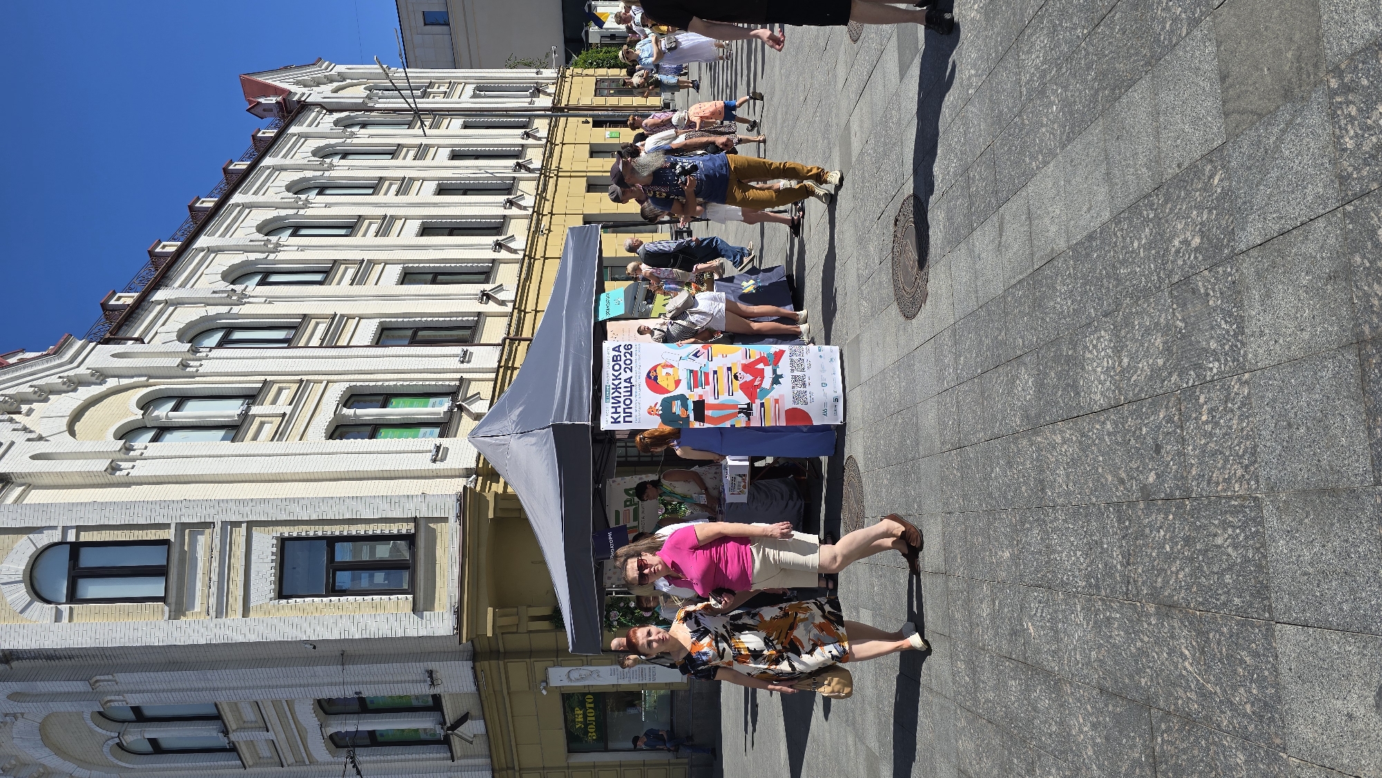 The image shows a street scene in Zhytomyr, Ukraine, likely during daylight hours due to the bright sunlight and clear blue sky. A large, multi-story building with ornate architectural details, painted yellow and white, dominates the background. In the foreground, a temporary stall with a gray canopy is set up, likely for a market or event. A large poster advertising "КНИЖКОВА ПЛОЩА 2026" (Book Square 2026) is prominently displayed at the stall. Several people are visible in the scene, browsing at the stall, walking along the street, or standing around. The ground is paved with large gray stone tiles. The overall impression is that of a public gathering in an urban environment.