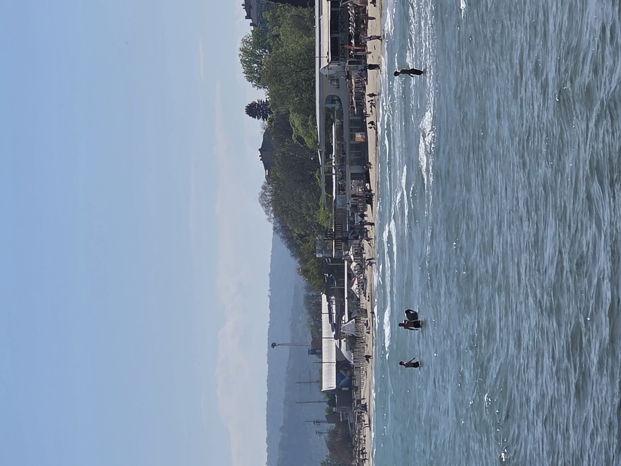 The image depicts a coastal scene in Varna, Bulgaria. A sandy beach lined with various structures, including buildings and shaded areas, runs along the shoreline. People are visible on the beach and in the water. The water appears choppy with visible wave patterns. Lush greenery covers the hillside that slopes down towards the beach. In the sky, blue suggests fair weather. The image is oriented vertically.