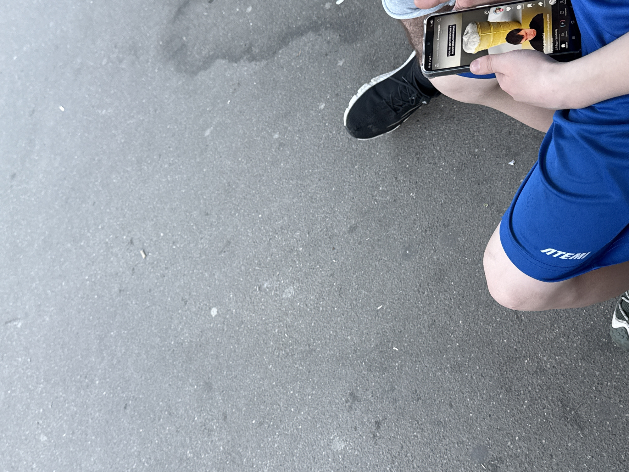 An overhead shot reveals a person sitting in an urban area, presumably in Moscow, Russia, looking at their smartphone. The phone screen displays a TikTok video. The individual is wearing blue athletic shorts with the brand name "ATEMI" visible on the left leg, and has black sneakers with white soles. A portion of another person's leg, clad in grey shorts, is also visible in the upper right of the frame. The ground surface is grey asphalt.