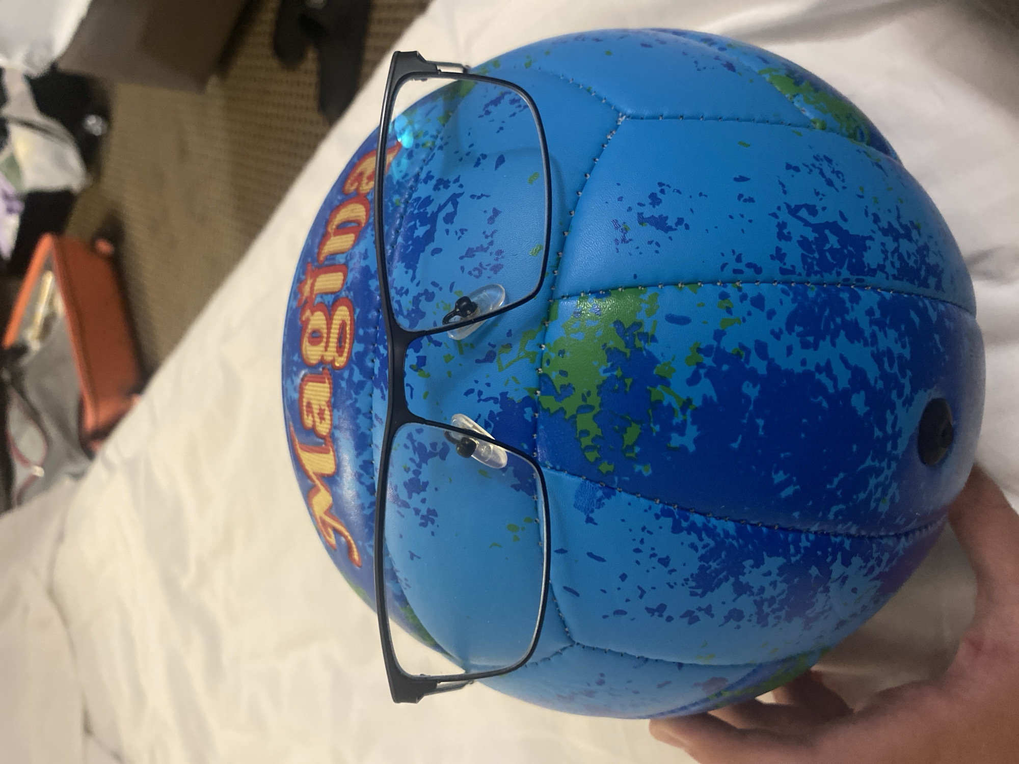 A blue volleyball with a world map print is positioned upright on a white, textured surface, likely a bed. The ball has a word printed on its upper hemisphere, partially visible as "Magina". A pair of black-rimmed glasses with clear lenses is placed on the ball, resting on the nose pads, giving the impression of a face. A hand is visible on the left side of the image, holding the ball. In the background, blurred out, are elements of a room including what appears to be luggage and furniture, suggesting an indoor setting, possibly a hotel room. The image was taken in the city of Nesebar, Bulgaria.