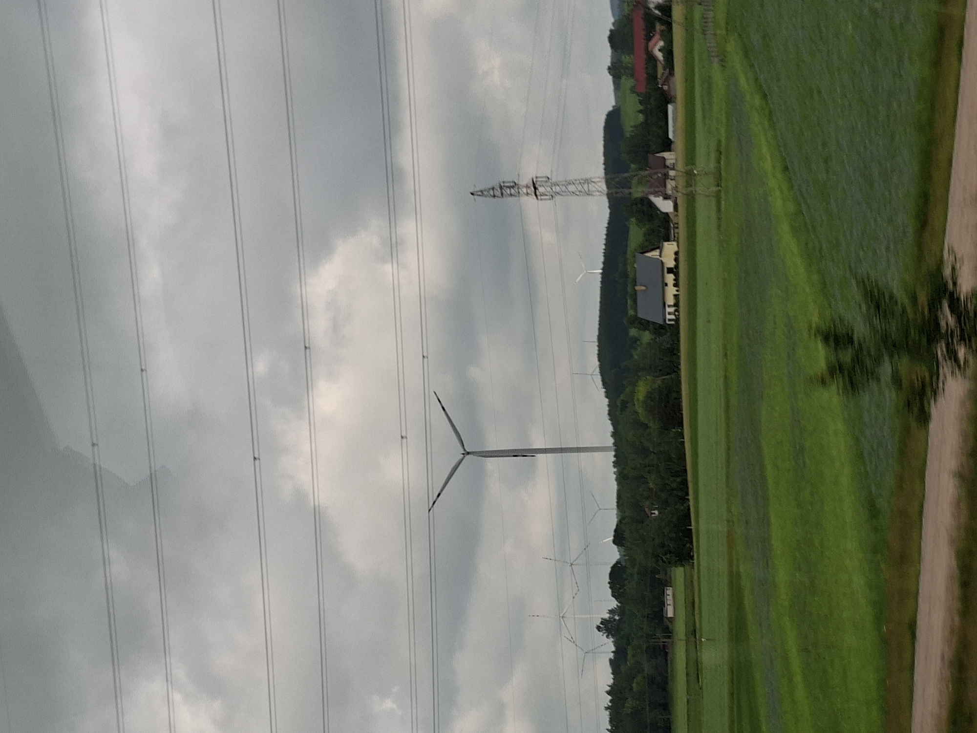 The image depicts a rural landscape under a cloudy sky. The main subjects are several wind turbines, some of which are in motion, suggesting they are generating electricity. The scene is a large, green field, possibly agricultural, with some darker patches that could indicate moisture or differing vegetation. In the background, a line of trees separates the field from a cluster of buildings, including houses with red and yellow roofs, and what appears to be a larger industrial structure, possibly a power pylon. The overall impression is one of a working landscape, focused on renewable energy production within a traditional rural setting. The weather appears overcast, typical of a cloudy day, and the time of day is indeterminate due to the lack of direct sunlight. No people are visible in the image, and there is no discernible text. The location context provided is Suwałki, Poland, which aligns with the presence of wind turbines and the rural architecture. The image is oriented vertically, making it a portrait-style photograph.