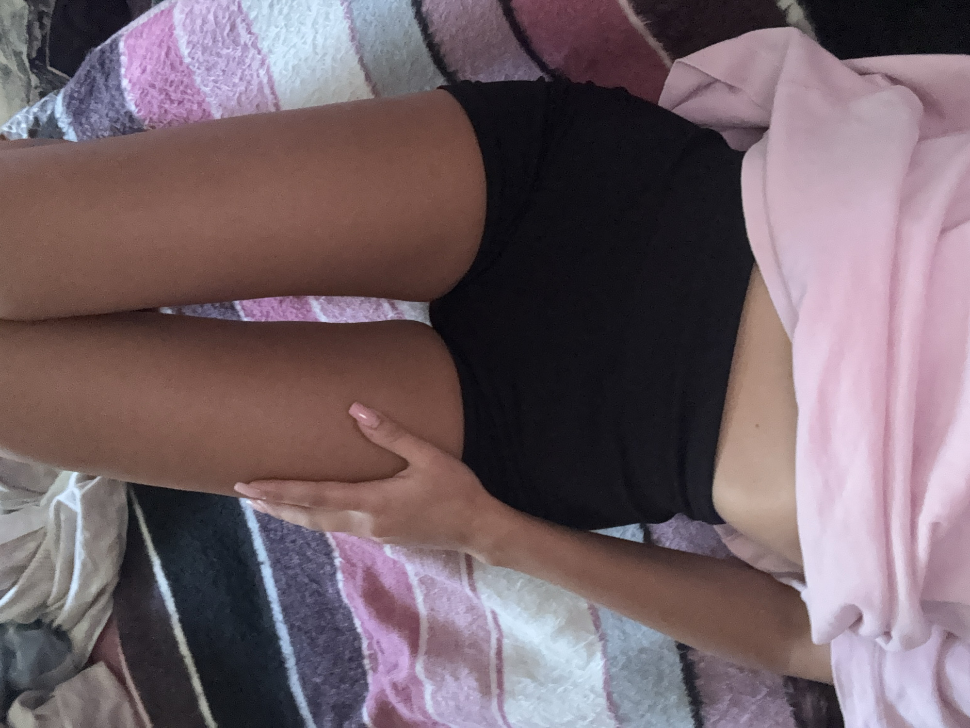 The image shows a close-up of a person's legs and torso, lying down on a striped blanket. The person is wearing black shorts and is partially covered by a pink blanket. The scene is likely indoors, given the blankets. The lighting suggests it could be daytime. The overall mood is relaxed and private. There is no visible text in the image. The location context of Basarabeasca, Moldova, does not provide any specific visual cues within the image itself.