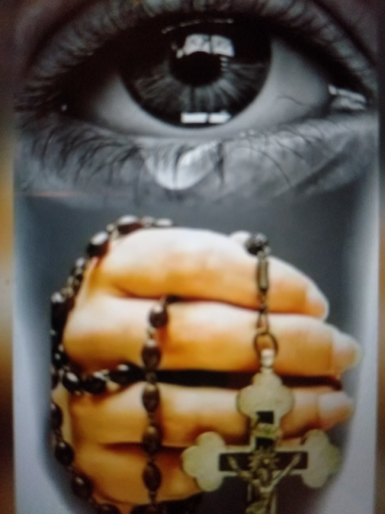 The image is a close-up, low-angle shot focusing on a person's eye and hand. The eye is dark with a tear visible on the cheek. The hand, appearing to be of a lighter skin tone, is clenched, and a rosary with dark beads and a crucifix is draped over it. The overall tone of the image is somber and suggests an emotional or spiritual moment. The background is blurred and neutral, drawing attention to the central elements. There is no visible text in the image. The location context provided is Jalingo, Nigeria.